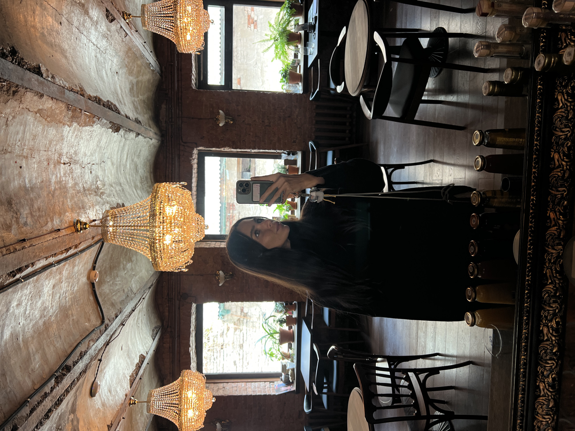 A woman in a black top stands in front of a framed mirror, holding a smartphone in her left hand, presumably taking a selfie. The reflection in the mirror shows a restaurant interior in Vladivostok, Russia, characterized by exposed brick walls, large windows with potted plants on the sills, and a vaulted ceiling adorned with crystal chandeliers. There are tables and chairs set up, and bottled beverages are arranged on shelves. The lighting is warm and ambient.