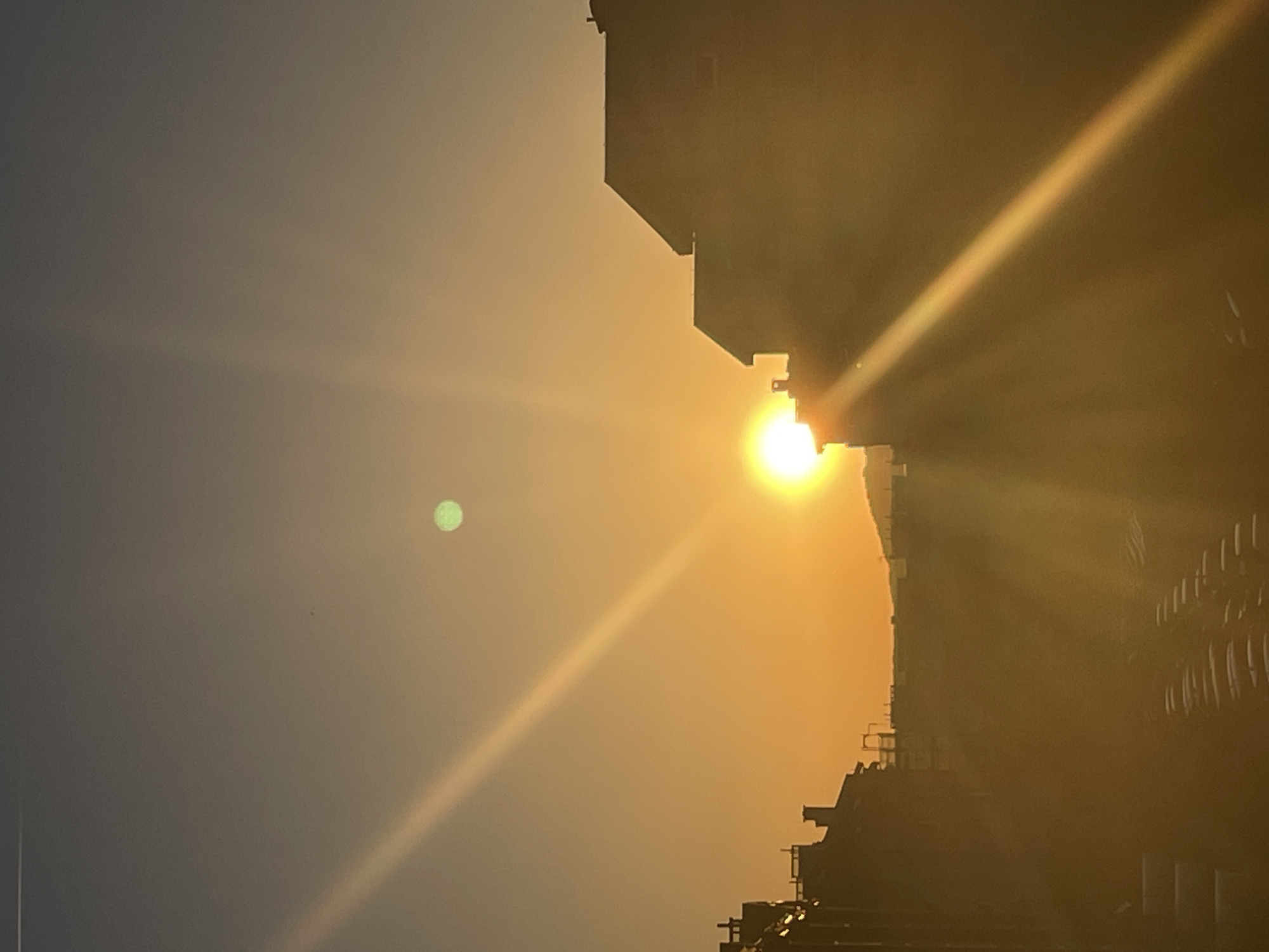 Here's a description of the image:
The image depicts a cityscape at sunset or sunrise. The sun is positioned to the right, partially obscured by the silhouette of a tall building. A bright beam of light extends diagonally from the sun across the frame. To the left of the sun, and seemingly in the sky, is a small, green orb. The sky transitions from a light orange near the horizon to a greyish-blue further up. The silhouette of the building is detailed with elements suggesting construction scaffolding or architectural features. The location is noted as city of Unknown, Unknown, offering no information about the location.
