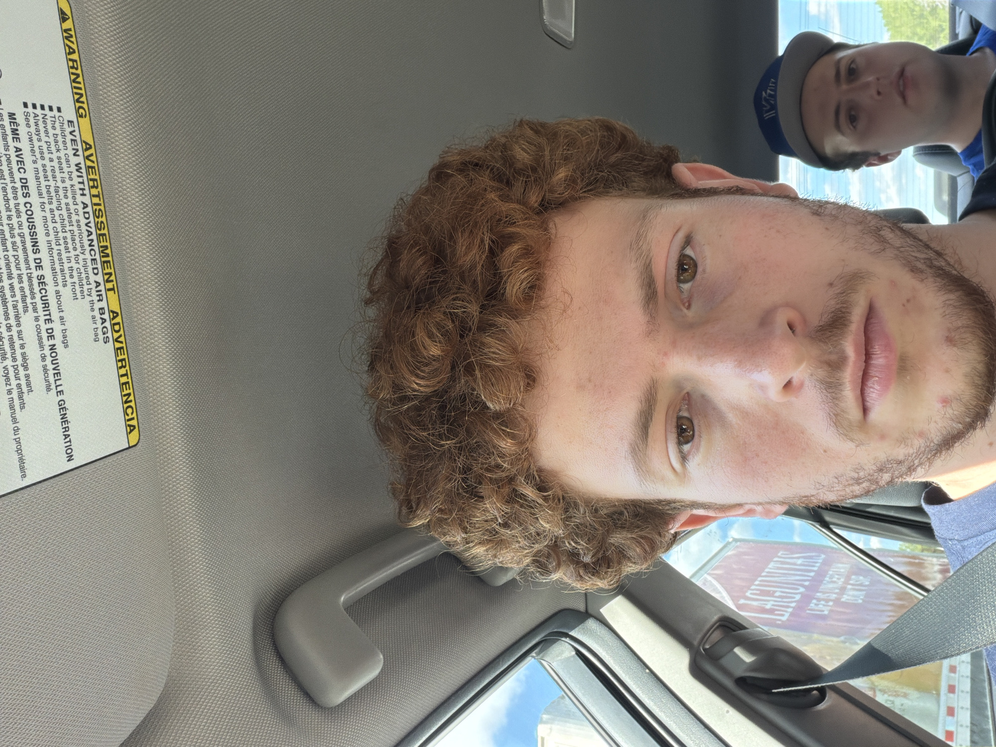 The image captures two males inside a vehicle on a bright, sunny day. The primary subject, a young man with light skin, curly reddish-brown hair, light brown eyes, and light facial hair, is prominent in the foreground. He wears a light blue shirt and has a neutral expression while looking directly at the camera. A seatbelt is visible across his chest.
In the background, a second male is partially visible, wearing a blue baseball cap and a blue shirt, also looking towards the front. The car's interior includes a light grey headliner, a grab handle, and a sun visor. Bright blue sky is visible through the car windows.
A prominent warning label on the sun visor is visible, containing text in English, French, and Spanish. The English portion reads: "AWARNING AVERTISSEMENT ADVERTENCIA EVEN WITH ADVANCED AIR BAGS", followed by bullet points: "Children can be killed or seriously injured by the air bag", "The back seat is the safest place for children", "Never put a rear-facing child seat in the front", "Always use seat belts and child restraints", and "See owner's manual for more information about air bags". French text, "MÊME AVEC DES COUSSINS DE SÉCURITÉ DE NOUVELLE GÉNÉRATION", is also present.
Outside the car window, a large red sign is partially visible, displaying the text "LAGUNITAS" and the slogan "LIFE IS UNCERTAIN DON'T SIP". The scene suggests the vehicle is in motion or parked in an outdoor setting during daytime in Teaneck Township, United States of America.