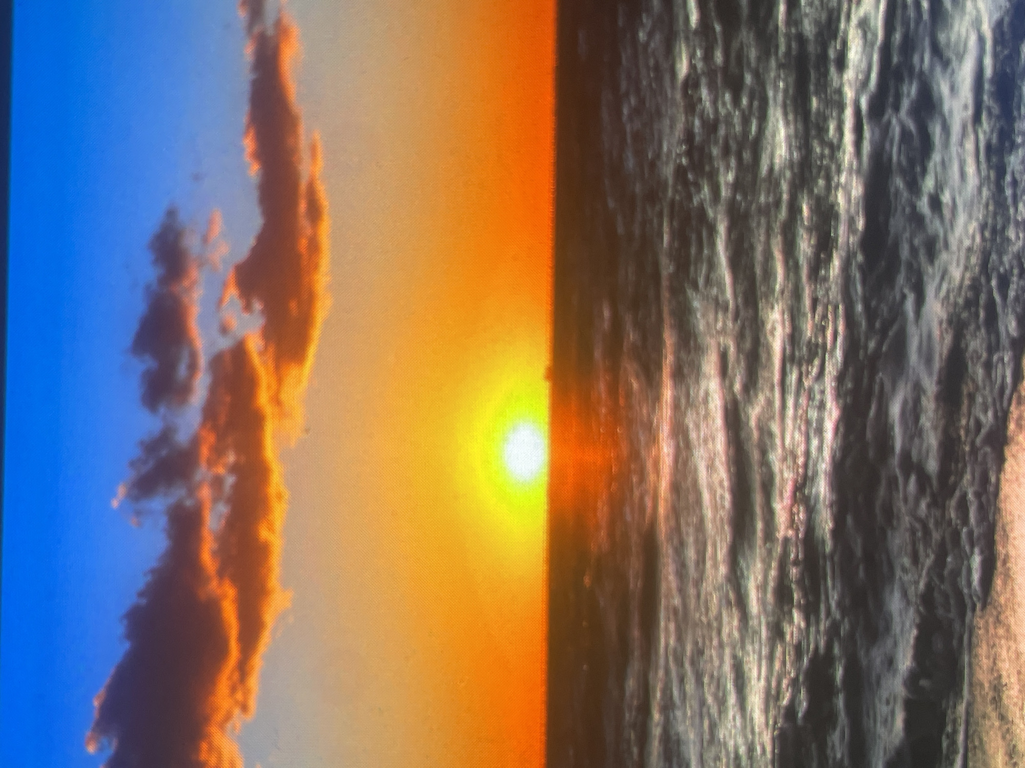 The image displays a sunset over a body of water, positioned at the horizon. The sky is divided into distinct color gradients: the upper portion is a vibrant blue, transitioning downwards into a bright orange and intense yellow directly surrounding the sun. Several clouds are present in the upper-mid sky, characterized by dark undersides illuminated by the orange sun, creating a contrast with their darker upper sections. The sun itself is a brilliant, circular, bright white and yellow disc situated precisely on the horizon line, casting a strong glow across the scene. Below the horizon, the water appears dark with noticeable wave patterns and ripples, reflecting a mix of the sky's orange and the ambient dark blue/grey, indicating movement on the surface. No people, subjects, objects, actions, or interactions are discernible within the frame. The environment depicted is a natural seascape. This scene is located in Zelenyi Hai, Ukraine.