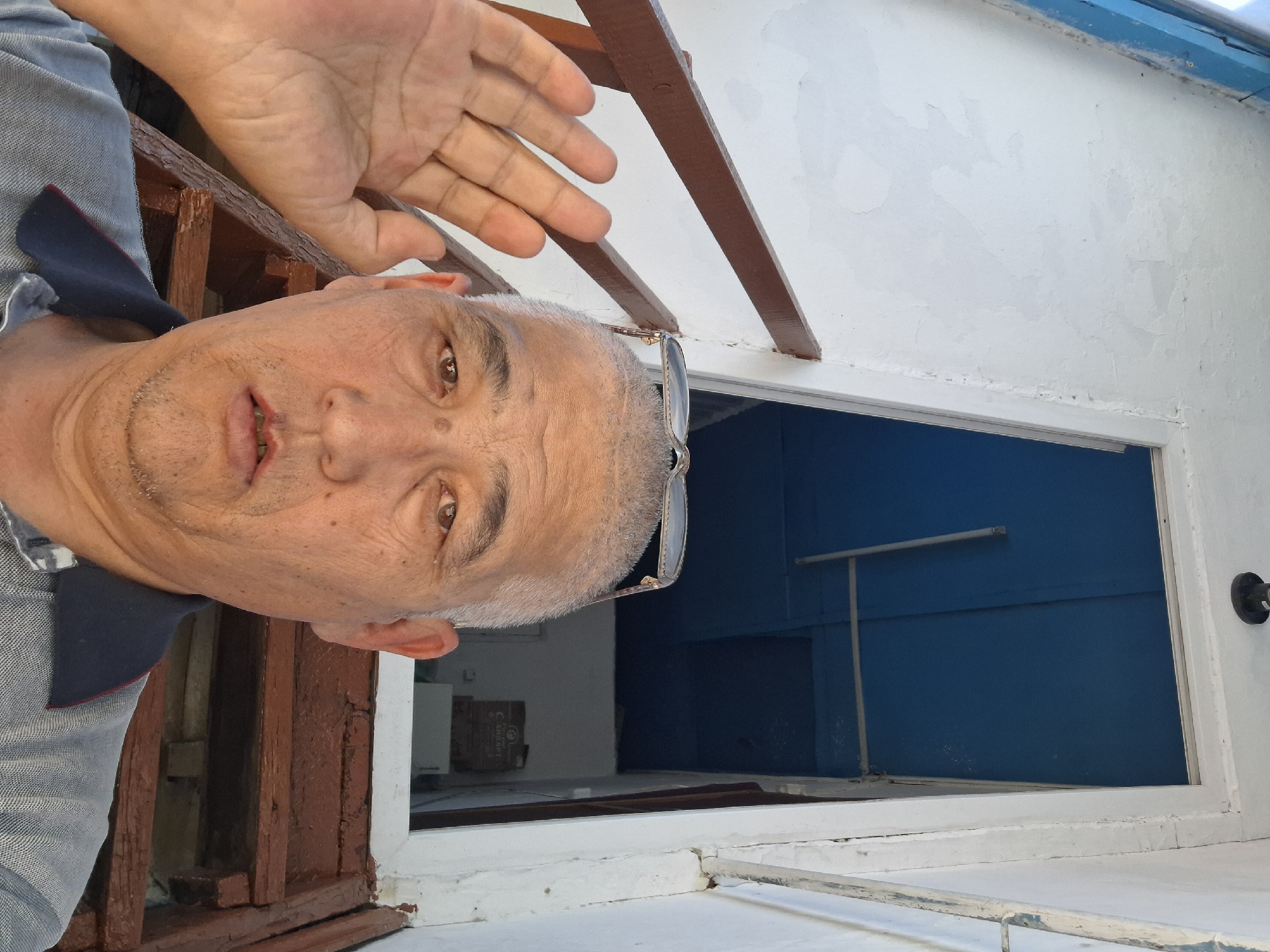 A close-up, selfie-style, eye-level shot features an adult East Asian male in the foreground, positioned slightly off-center to the right. He has short, graying hair, weathered skin, and appears to be wearing a gray collared shirt. His right hand is raised, palm facing the camera, as if waving or gesturing. He is looking directly at the camera with a neutral expression. On top of his head, resting on the back of his head, is a pair of eyeglasses.
The background reveals a weathered wooden staircase with dark brown railings leading upwards. The staircase is adjacent to a white building with peeling paint. Beyond the staircase, an open doorway leads into a space with a bright blue ceiling and walls. Visible through the doorway is a glimpse of what appears to be an indoor area with some boxes and possibly a white appliance.
The scene suggests an outdoor or semi-outdoor location, possibly residential. Without further context or distinctive landmarks, pinpointing an exact location within Almaty, Kazakhstan is not possible. The quality of the building and stairs suggests an older or less maintained structure. The lighting indicates it is daytime, likely sunny.