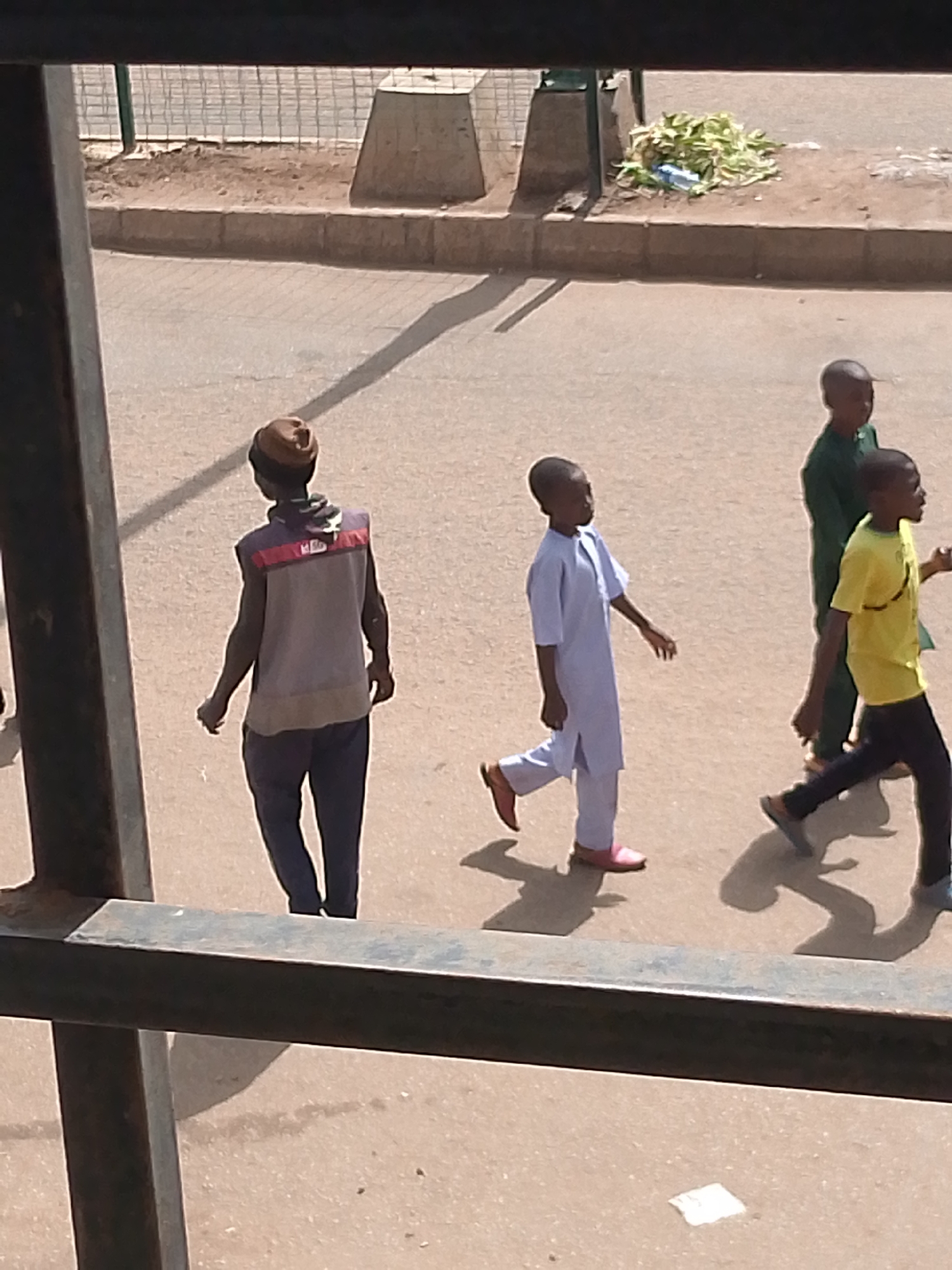 The image shows a street scene in Bukuru, Nigeria, with four people walking. In the foreground, a person wearing a brown cap and a sleeveless vest with red and grey stripes walks away from the camera. Behind them, three boys are walking in the same direction. The boy closest to the camera wears a light blue traditional outfit and pink shoes. The boy next to him wears a yellow t-shirt and dark trousers. The farthest boy is only partially visible, wearing a green shirt and dark trousers. They are walking on a paved surface, likely a road or sidewalk, with shadows indicating it is daytime, likely sunny. In the background, there is a curb, a pile of green vegetation and a blue object, possibly a water bottle, and some concrete structures. The overall scene appears to be a normal street activity, with no particular emotions or event being conveyed. There is no visible text in the image.