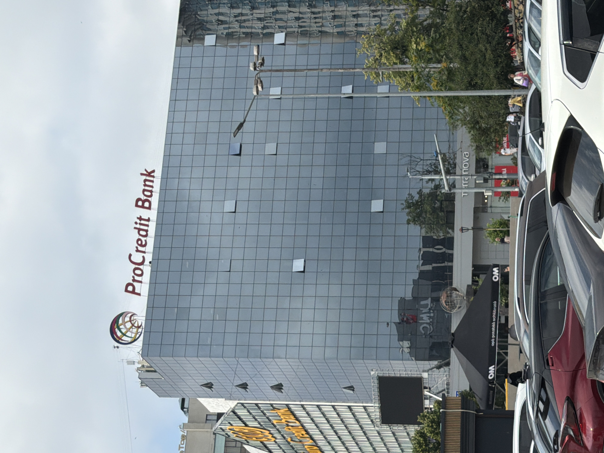 The image shows the ProCredit Bank building in Chișinău, Moldova. The building is a modern glass-facade structure with the ProCredit Bank logo prominently displayed at the top. In front of the building, a parking lot is visible with several cars. The sky is overcast with clouds. A large black umbrella with "OM" logo and "apa potabilă echilibrată" text is set up near the entrance of the building, suggesting a commercial setup. In the background, other buildings and trees are visible. A street lamp stands in front of the glass building. The overall scene depicts an urban environment with a focus on the ProCredit Bank building and its surroundings.