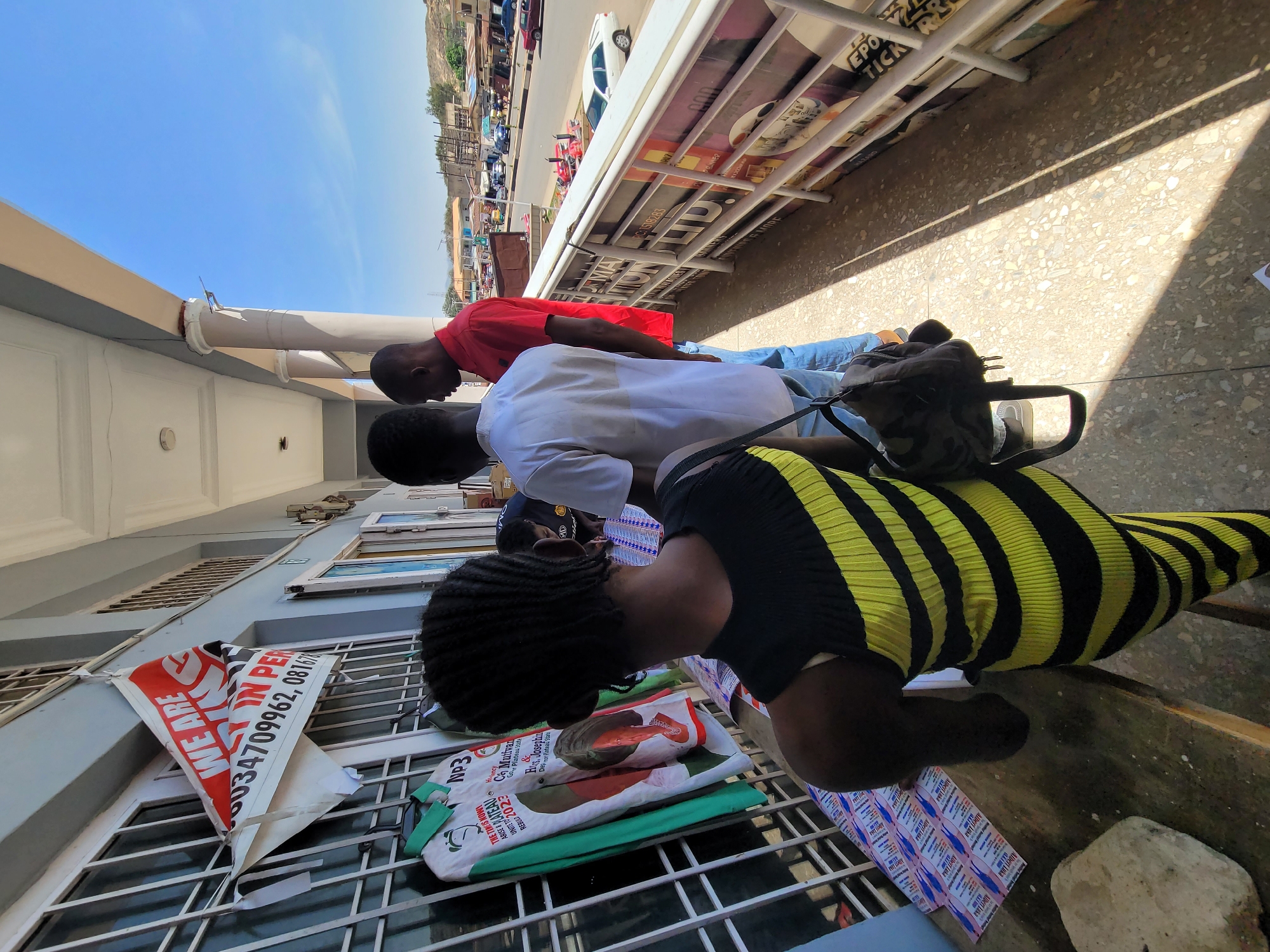 The image depicts an outdoor urban scene in Jos, Nigeria, under bright sunlight, suggesting midday or early afternoon due to the clear blue sky and strong shadows. The setting appears to be a commercial or public area, with a light-colored building facade featuring windows and grates on the left, and a street with vehicles and buildings on a distant hillside in the background.
Four individuals are visible. In the foreground, a person with dark braided hair, dressed in a black and yellow horizontally striped garment, leans forward, looking down at items on a gridded surface. Behind them, two men stand: one in a white short-sleeved shirt and blue jeans, with a dark bag slung over his shoulder, and another in a red short-sleeved shirt and blue jeans. Both are looking forward or down. A fourth person's head is partially visible further back.
Various objects are present, including a camouflage-patterned bag on the ground. Prominently displayed are several items with text, indicating a political context. A red and white banner attached to the building reads "WE ARE IN PER" with phone numbers "6034709962, 081678". On the surface in front of the bending person are green and white materials, likely campaign bags or flyers, bearing slogans such as "THE TIME IS NOW", "Np3", "ARISE & REBUILD 2023", "C Mutfwan", "Go Plateau State", and "& H Josephin Der mor Plateau Stat". These texts strongly suggest election campaign materials for Plateau State in 2023. Small flyers are also scattered on the ground. A large white sign with text like "RIZ", "PC:1518283", and "EPO" is visible in the background. The activity suggests people are engaging with or examining these displayed materials, possibly at a political campaign point or public information stall.