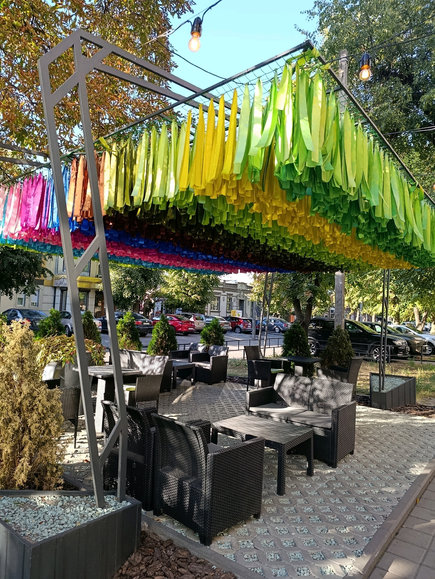 The image depicts an outdoor seating area of a cafe in Cherkasy, Ukraine. A metal canopy structure is adorned with numerous colorful ribbons, predominantly in shades of green and yellow, with accents of pink, blue, and orange visible towards the left side. These ribbons hang in dense rows, creating a vibrant, ceiling-like effect. Beneath the canopy, several tables and chairs made of dark woven material are arranged on a paved surface composed of hexagonal concrete slabs filled with small white pebbles. In the foreground, a dark planter box filled with gravel and a sparse, dry-looking shrub is visible. Beyond the seating area, trees line a street where cars are parked, and buildings can be seen in the background. Two outdoor light bulbs hang from wires above the canopy. The scene is bathed in natural daylight, suggesting it was taken during the day. There are no people visible in the image.