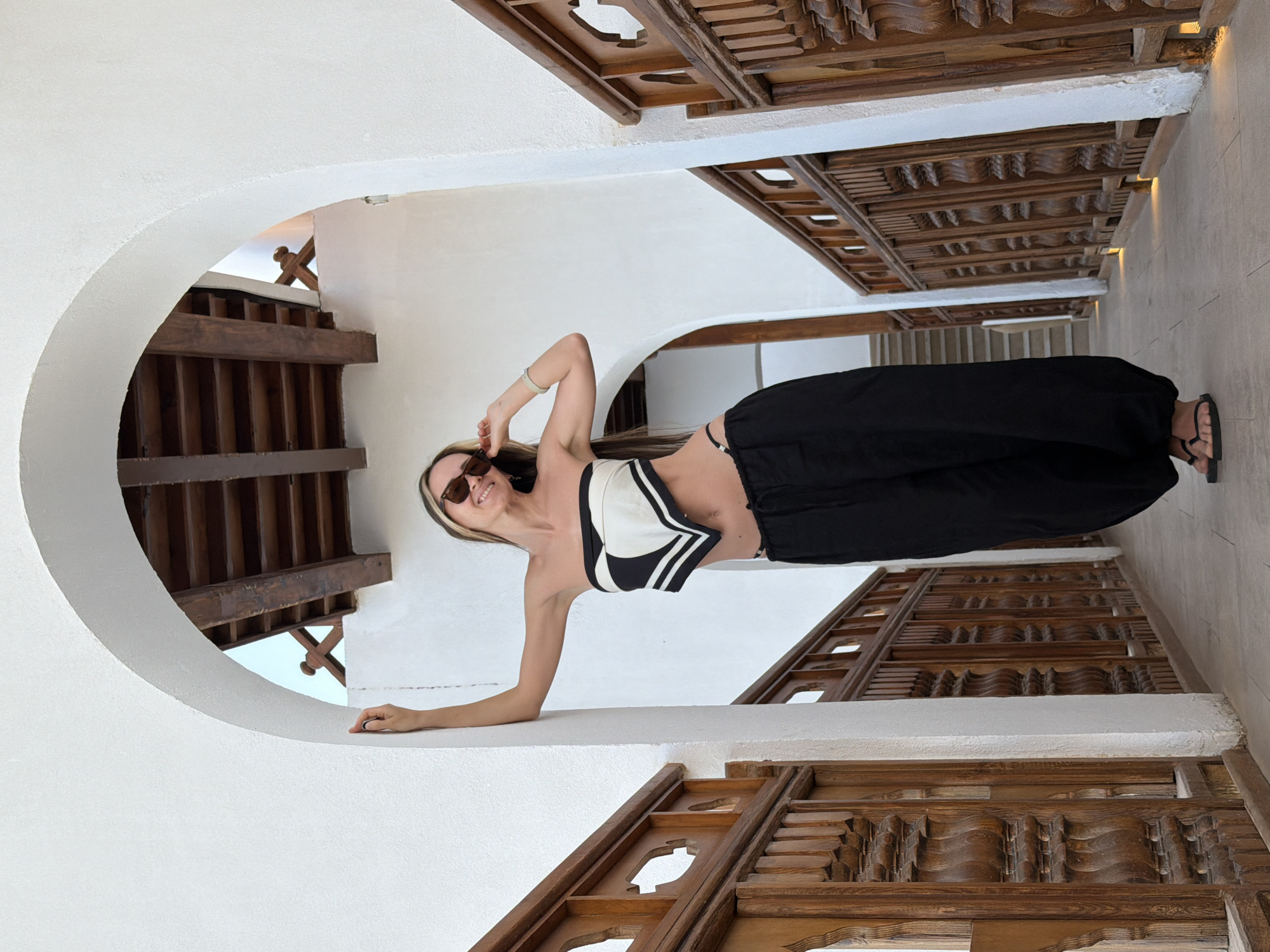 A woman wearing a black and white top, black pants, and sunglasses is posing inside an arched window in what appears to be a building in Sharm El Sheikh, Egypt. The arched window is white, and a wooden decorative structure is visible on either side. A staircase is visible in the background.