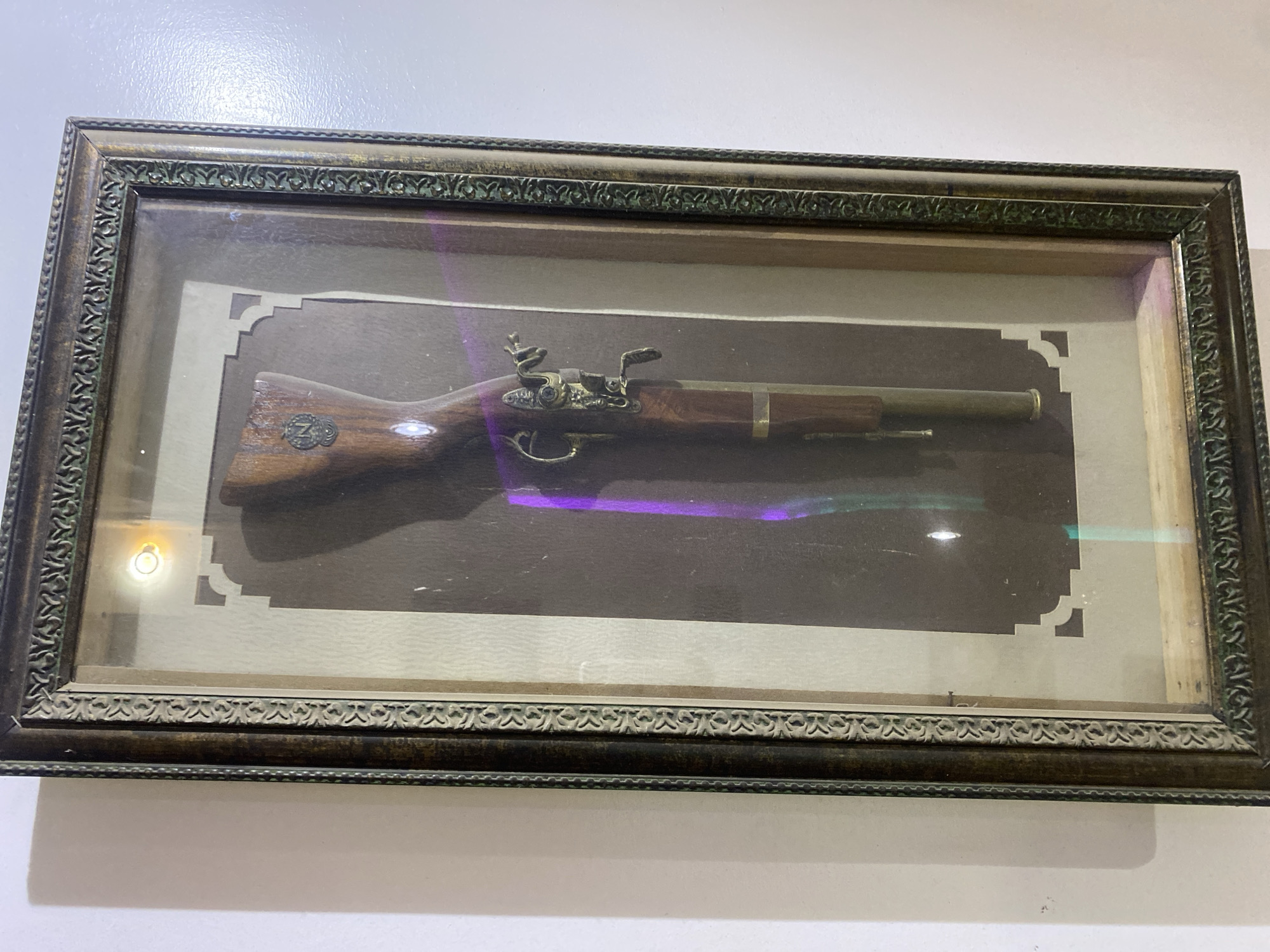 A framed antique flintlock pistol is displayed on a wall in Giginyu, Nigeria. The pistol, with a wooden stock and brass hardware, is mounted on a dark brown backing with an off-white decorative border. The frame is ornate and dark brown with gold accents. The lighting in the image suggests an indoor setting, possibly a home or display case. There are no people visible in the image.