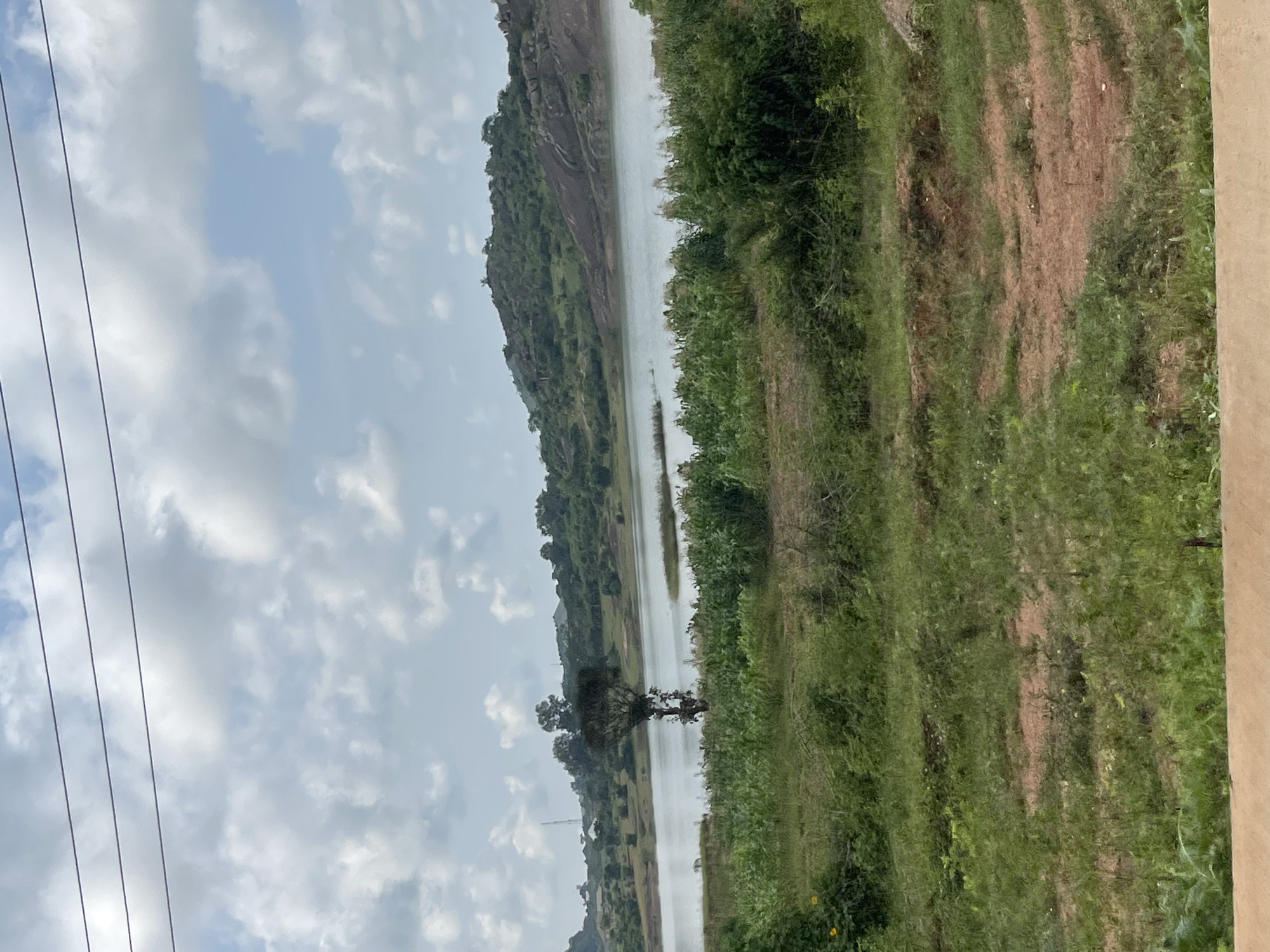 The image depicts a landscape view of a body of water, likely a reservoir or lake, situated in the city of Jos, Nigeria. In the foreground, there is a mix of green vegetation and reddish-brown earth. The middle ground features lush green foliage, including what appears to be a field of crops, possibly corn, bordering the water. A prominent, solitary tree stands near the water's edge. The background reveals a hilly terrain covered with green vegetation and rocky outcrops. The sky is partly cloudy with a light blue hue and scattered white clouds. There are overhead power lines visible in the upper portion of the image. No people or animals are present in the scene. The overall environment suggests a rural or semi-rural setting within Jos.