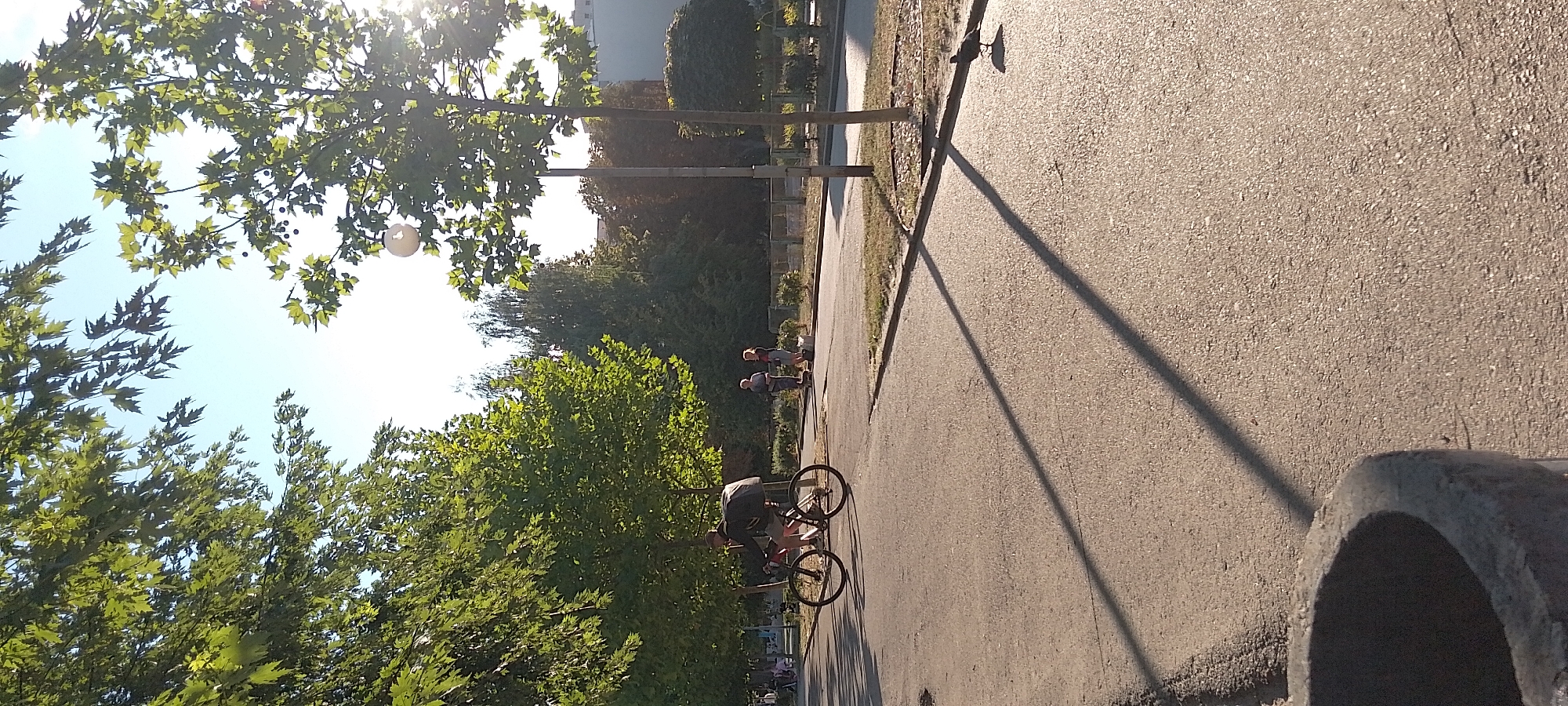 A cyclist rides along a paved path on a sunny day, casting a long shadow across the asphalt. The cyclist is wearing a dark top and shorts, and is carrying a large black bag on their back. The path is lined with trees and grass. In the background, two people are standing on scooters, and a bird is walking on the path. The scene appears to be in a park or recreational area within the city of Cherkasy, Ukraine, possibly near a residential area, given the presence of buildings in the distance. The lighting suggests it is late afternoon or early evening.