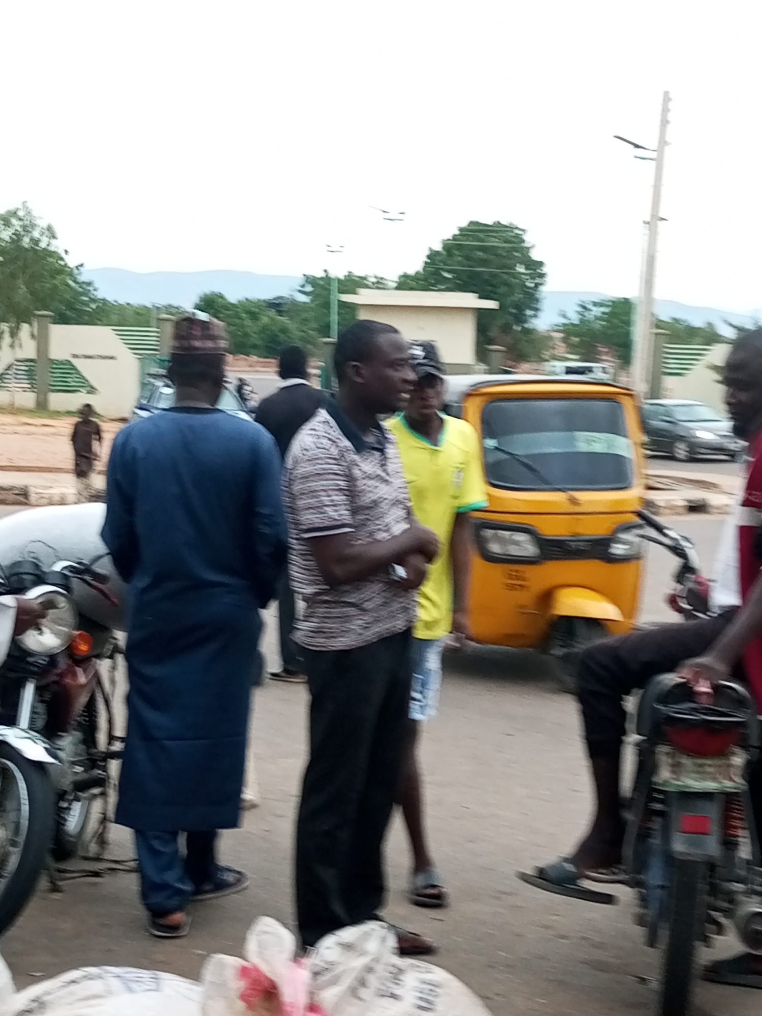 The image depicts a street scene in Gombe, Nigeria, likely during daylight hours, with a mix of individuals, vehicles, and buildings. In the foreground, a person in a blue garment stands with their back to the camera, facing a yellow auto-rickshaw. To the right of the auto-rickshaw, another individual wearing a red and white shirt is partially visible. Several other people are scattered throughout the scene, some standing and others in motion.
The environment includes a paved road, a sidewalk or dirt path, and some low-lying vegetation. In the background, buildings with light-colored walls and green trim are visible, possibly part of a market or commercial area. A utility pole with lights stands prominently on the right side of the image. A motorcycle is parked on the left, with its front wheel and headlight clearly visible. The overall atmosphere suggests a bustling, everyday urban environment.