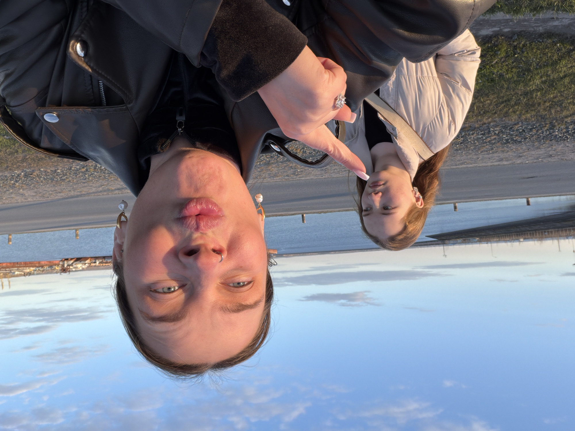 Two individuals are pictured in an outdoor environment in Chaikovsky, Russia, with the sky and a body of water visible in the background. The person on the left has their face closer to the camera, wearing a black leather jacket, with fair skin, light eyes, and jewelry in the ear and nose. This individual is making a kissing face. The person on the right is wearing a light-colored puffy jacket. One of the individuals is pointing at the other with a hand adorned with a large ring. The location appears to be near a port, with industrial structures visible on the horizon. The image is oriented upside down.