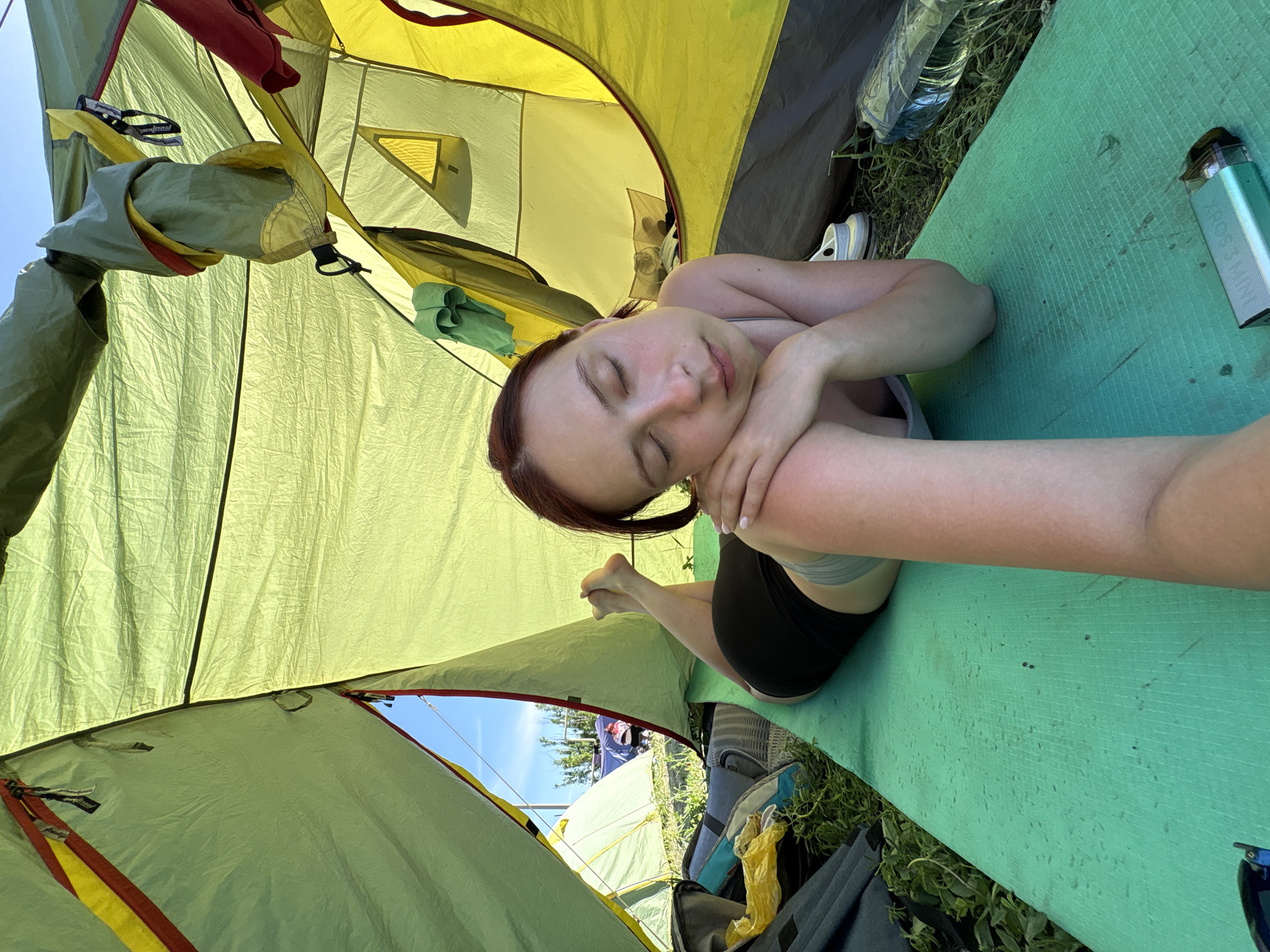The image shows a woman lying inside a tent. She has closed eyes and is resting on a green mat. She is wearing dark shorts and a light-colored top. The tent is yellow and green, with other tents visible outside. There is a water bottle, a pair of white shoes, and a small electronic device next to her on the mat. The location is likely in Khvalynsk, Russia, but there is no specific evidence within the image to confirm the exact location.