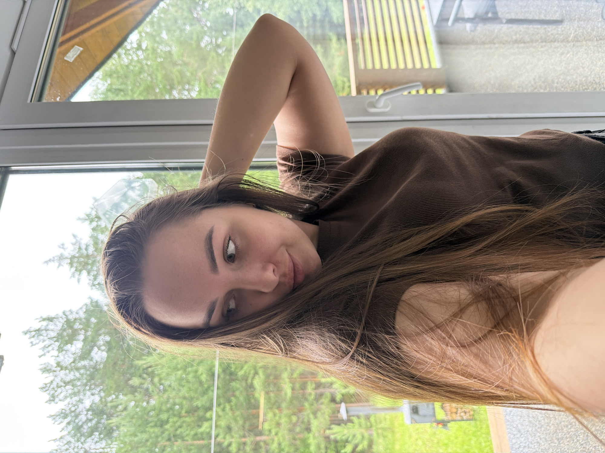 The image shows a young woman lying down indoors, with her arm raised above her head. She is wearing a brown long-sleeved shirt. Her hair is long and brown, falling around her face and shoulders. She is looking towards the camera with a relaxed and playful expression. The background appears to be a window overlooking a lush green outdoor setting, likely a yard or garden with trees. The lighting suggests it is daytime, possibly overcast due to the soft light. The scene conveys a sense of casual comfort and personal moment captured.