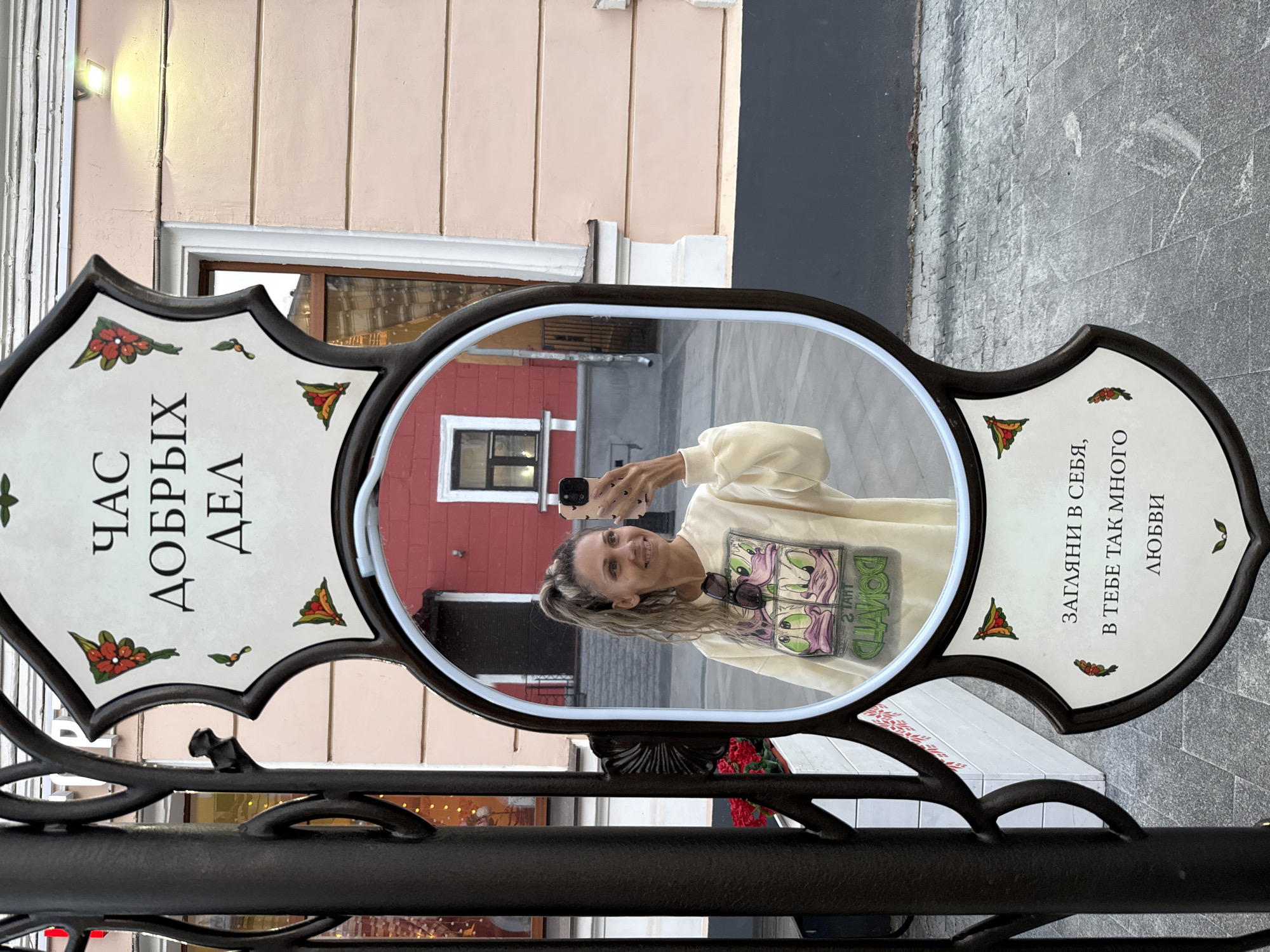 The image shows a woman taking a mirror selfie outdoors, with a decorative sign featuring Russian text and floral motifs. The sign is mounted on a black metal post, with a mirror in the center reflecting the woman and her surroundings. She is wearing a light-colored sweatshirt with a cartoon print. In the reflection, a red building with windows is visible behind her. The ground is paved with gray tiles. The sign has text that translates to "Time for good deeds" at the top and "Look into yourself, there is so much love in you" at the bottom. The location is identified as Rusanovo, Russia.
