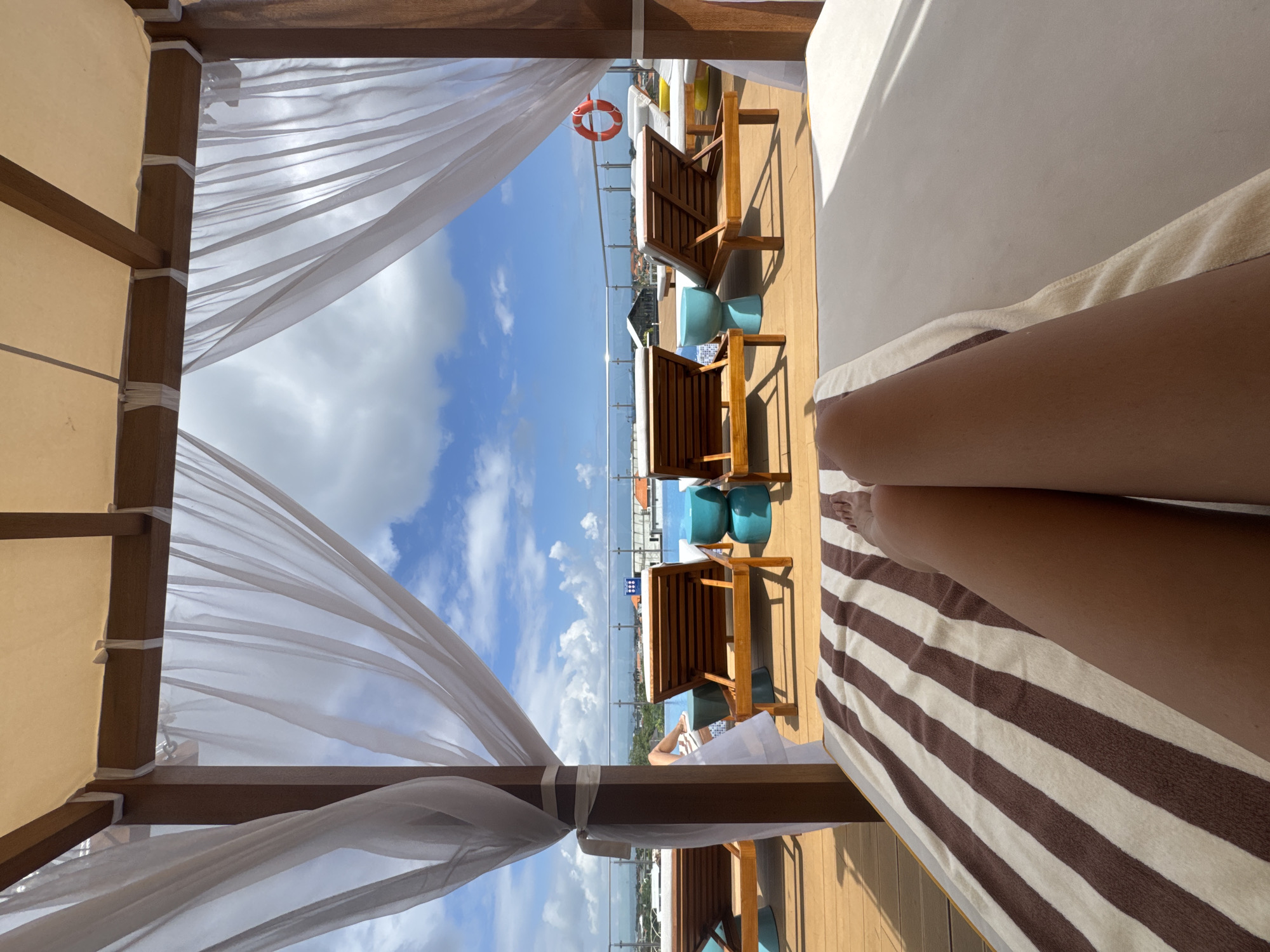 In North Kuta, Indonesia, an outdoor cabana provides shade over a sunbathing area. The cabana's roof is made of tan fabric stretched over a wooden frame, with sheer white curtains hanging from the sides. The foreground shows a person's legs resting on a lounge chair with a striped brown and white towel. Beyond the cabana, several empty wooden lounge chairs are arranged on a wooden deck around a bright blue swimming pool. In the background, a glass railing overlooks a cityscape with buildings and a cloudy blue sky. A life preserver is visible hanging on the railing. The scene suggests a resort or hotel setting.