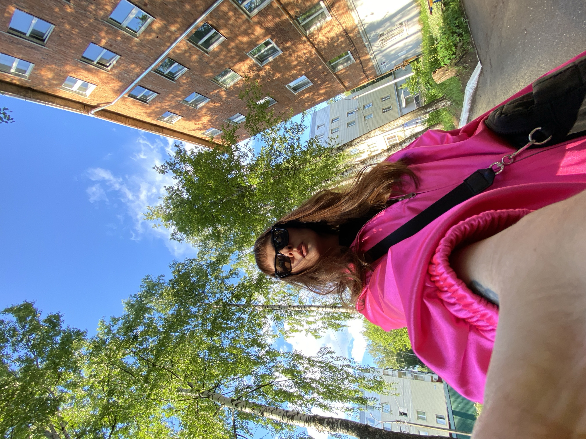 A woman wearing sunglasses and a bright pink jacket stands in Votkinsk, Russia. The image is taken from a low angle, looking up at the woman, the sky, trees, and the surrounding buildings. A red brick building and a light-colored apartment building are visible in the background. The sky is blue with scattered clouds.
