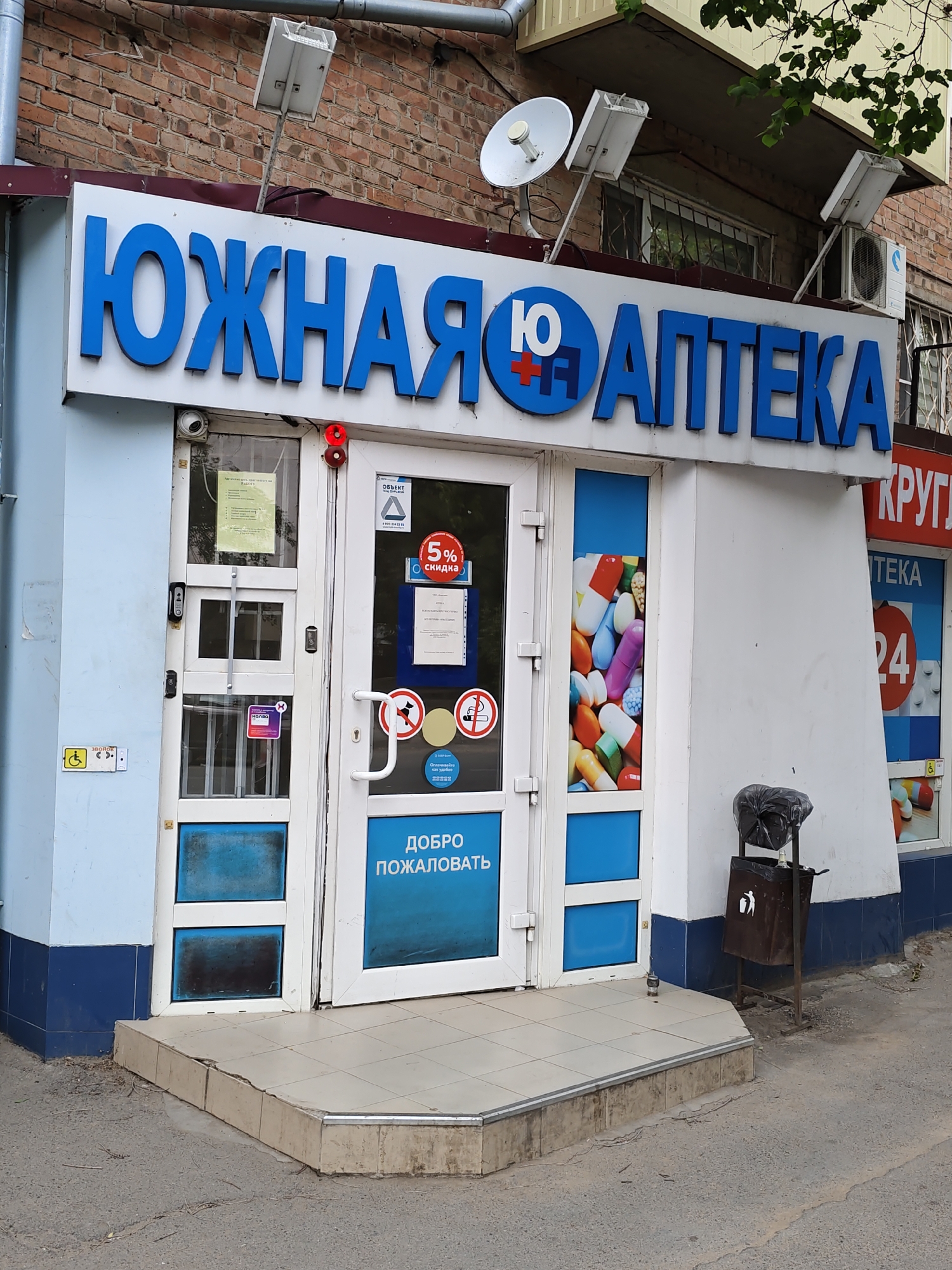 The image shows the exterior of a pharmacy in Rostov-on-Don, Russia. The pharmacy's sign reads "ЮЖНАЯ АПТЕКА" (YUZHNAYA APTEKA), which translates to "Southern Pharmacy." The entrance features double glass doors. To the left of the entrance, a blue sign with a wheelchair symbol indicates accessibility. Above the entrance, a security camera is visible. A red blinking light is mounted on the door frame. On the glass door, there are several stickers, including one that says "5% скидка" (5% discount) and another with a "no smoking" symbol. Inside the pharmacy, visible through the glass, is a display of colorful pills. To the right of the entrance, there is another pharmacy sign that says "КРУГЛ" (KRUGL), likely part of a larger sign for a 24-hour pharmacy. A trash can with a black trash bag is located next to the entrance. The building is made of brick, and there is a metal pipe and a satellite dish mounted on the facade. The ground in front of the pharmacy is paved with tiles, and there are some scattered leaves and debris.