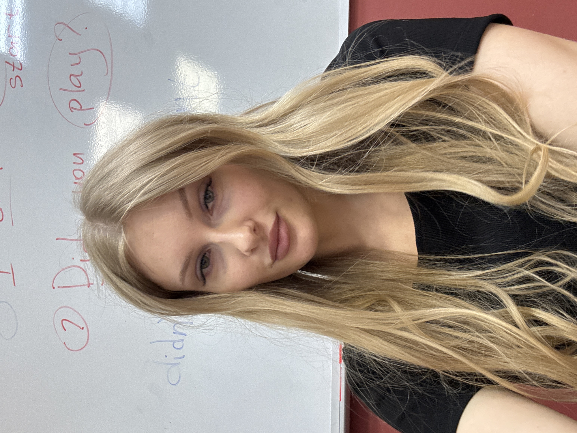 A young woman with long blonde hair is posing in front of a whiteboard. The whiteboard has handwritten notes in red and blue marker, including "Did you play?". The woman is wearing a black top. The location is likely indoors. I cannot verify if the location is in Perm, Russia.