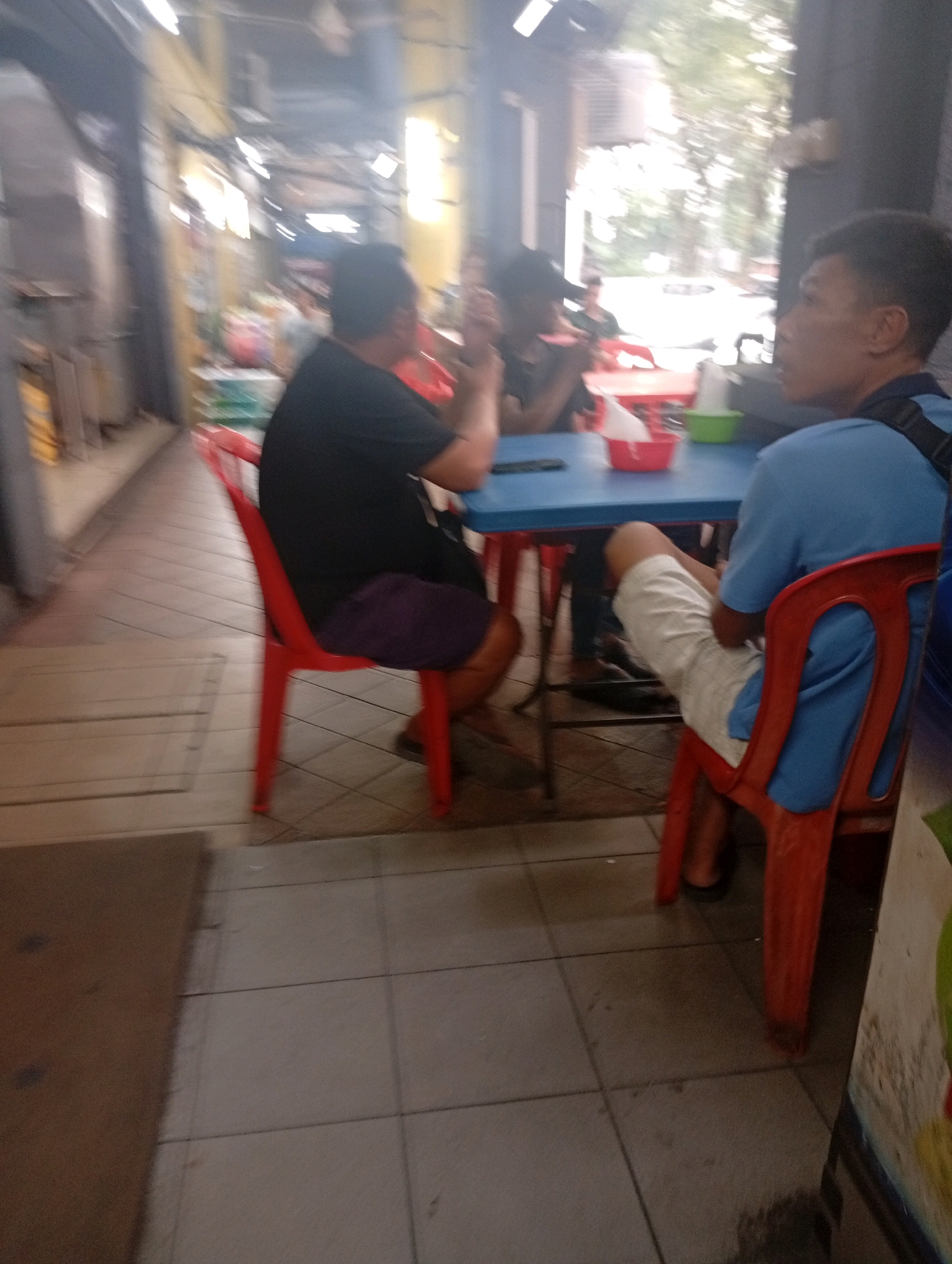 A group of people are sitting at a table in an outdoor food court setting in Selayang Municipal Council, Malaysia. Two men are visible in the foreground, one wearing a blue t-shirt and white shorts, and the other in a black t-shirt and dark shorts. They are seated at a blue table, with a red plastic basket containing what appears to be napkins or food wrappers on the table, along with a green bowl. The man in the blue shirt is looking to his right, and the man in the black shirt is looking forward. Another person, partially obscured, is seated to the right of the man in the black shirt. Red plastic chairs are present, with one occupied by the man in the black shirt. The environment appears to be a covered walkway or alleyway, with tiled flooring and overhead lighting fixtures. The background is blurred, but suggests trees and other structures, consistent with an urban outdoor dining area.