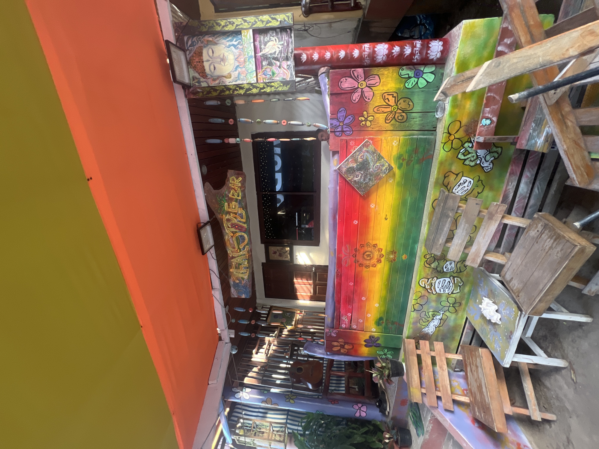 The image shows the exterior of a brightly painted bar called "THAI STYLE BAR" in Ban Tai Subdistrict, Thailand. The bar features a rainbow-colored facade adorned with floral motifs and other artwork, including a depiction of Buddha. Several wooden chairs and tables are arranged in front of the bar. A guitar is visible hanging on a rack behind the bar, and plants are placed on steps leading up to the entrance. The area is covered by an orange and yellow awning, and outdoor lighting fixtures are mounted above the bar. The scene appears to be outdoors, with sunlight casting shadows.