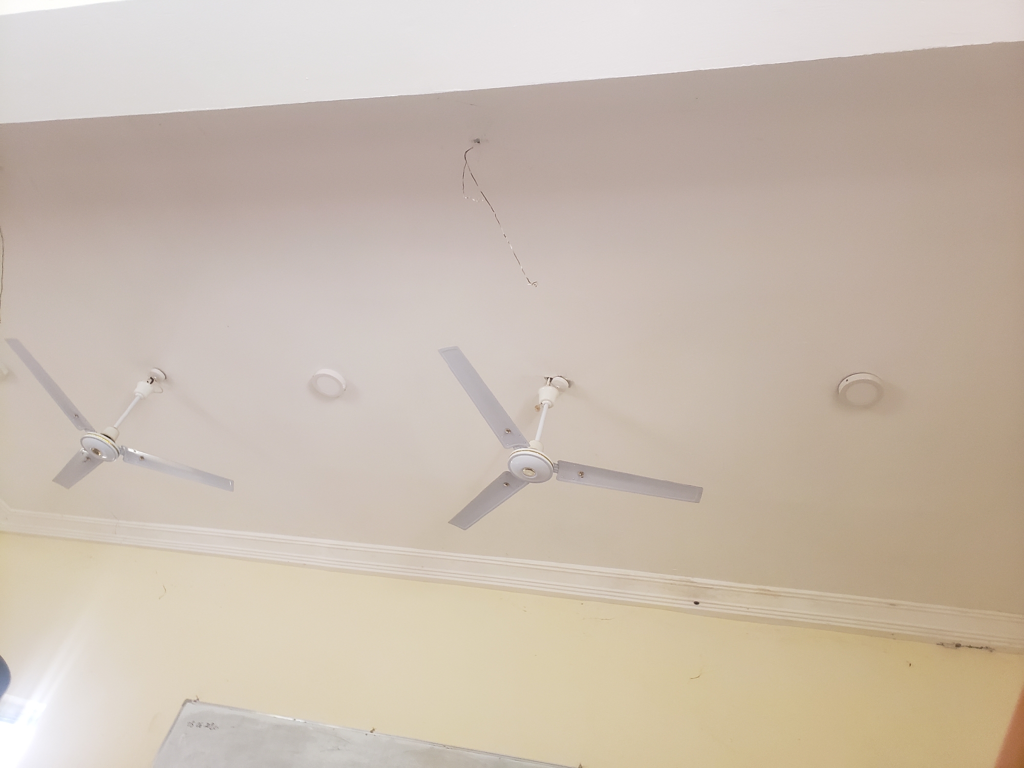 The image shows the interior of a room with a white, textured ceiling and light yellow walls. There are two white ceiling fans visible. One is on the left side of the image, and the other is to the right of the center. Both fans are suspended from the ceiling by white rods and have three white blades. Above the left fan, a small, circular white object is attached to the ceiling. To the right of the center fan, a similar circular white object is also attached to the ceiling, possibly a smoke detector or light fixture. A thin, metallic wire hangs down from the ceiling near the top center of the image. The wall below the ceiling has a decorative molding running horizontally. A portion of a light-colored surface, possibly a whiteboard or projection screen, is visible in the lower left corner, with some faint markings on it. The lighting in the room appears to be coming from an unseen source, casting a soft illumination. The image is taken from a low angle, looking upwards towards the ceiling. The location is identified as Gumel, Nigeria.