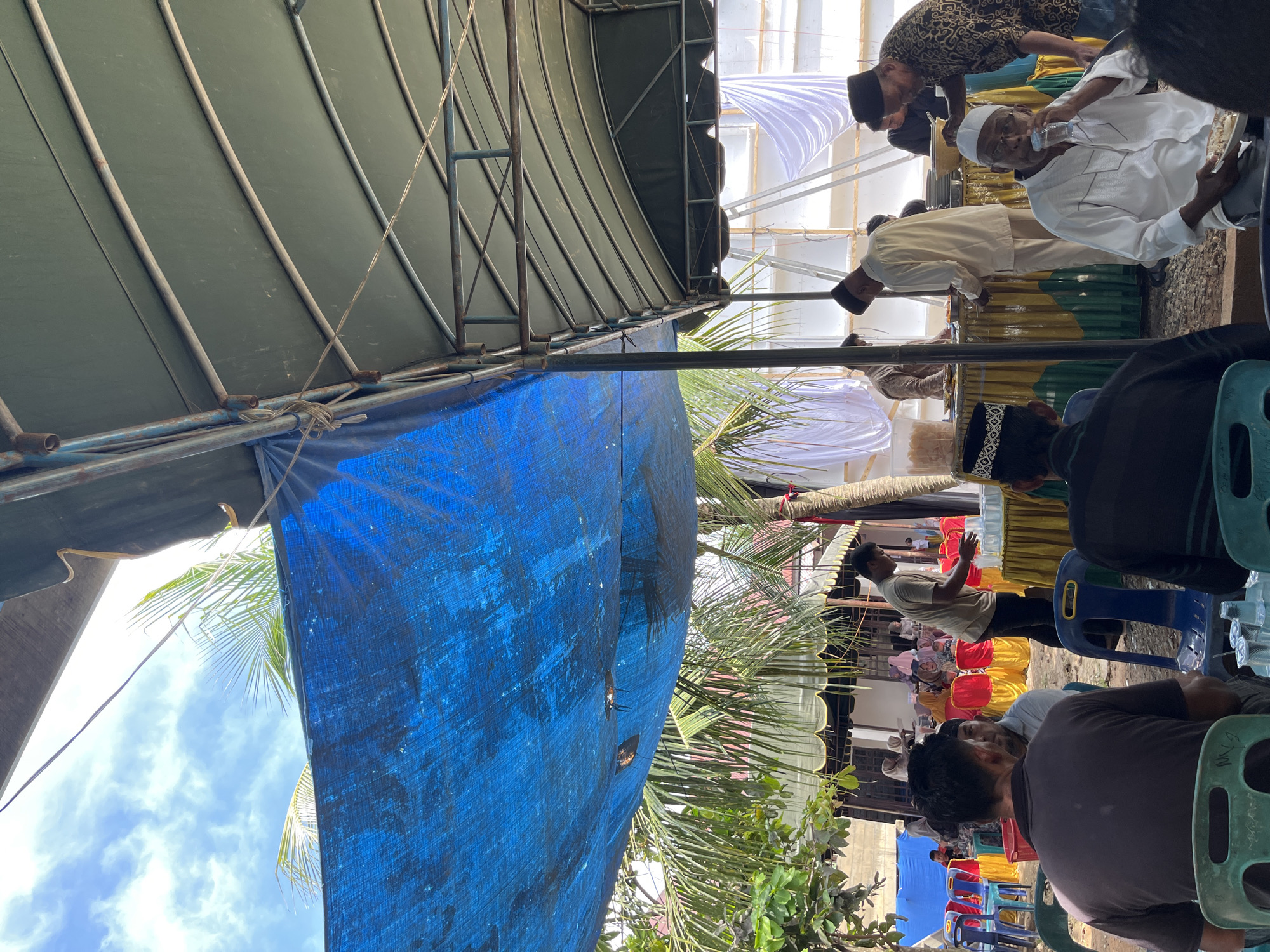 This image captures an outdoor gathering under temporary shelters in Manyang, Indonesia, likely during a daytime event.
In the foreground, two men are seated with their backs to the viewer on green plastic chairs. To their right, another man, wearing a white shirt and a white peci, is seated on a blue plastic chair, drinking from a clear glass with a plate of food in his left hand. Directly behind him, a man in a patterned long-sleeved shirt and black peci stands next to a table covered with a yellow and green striped cloth, appearing to manage items on it. To the left of this table, another man in a light-colored long-sleeved shirt is attending to items on a similar table. Further back, under the green-canopied tent, more individuals are visible, some serving, others standing near food service areas.
The scene is dominated by two primary overhead coverings: a large blue tarp, visible on the left and partially across the top, and a green-colored canopy tent on the right, supported by a metal frame structure. Sunlight is bright, casting shadows and illuminating parts of the scene, particularly the sky visible through an opening in the blue tarp, revealing scattered clouds. Palm trees and other green foliage are prominent, with palm fronds extending into the frame, especially on the left.
In the background, under what appears to be another tent or structure, a larger group of people, including women wearing headscarves (hijabs), are seated at tables with red and yellow chairs. White fabric or tarps are used as backdrops or walls for a structure further in the distance. The ground appears to be dirt or uneven terrain. Scattered plastic cups and other small items are visible on tables and on the ground. The overall impression is an informal community gathering or celebration with shared food and drink.