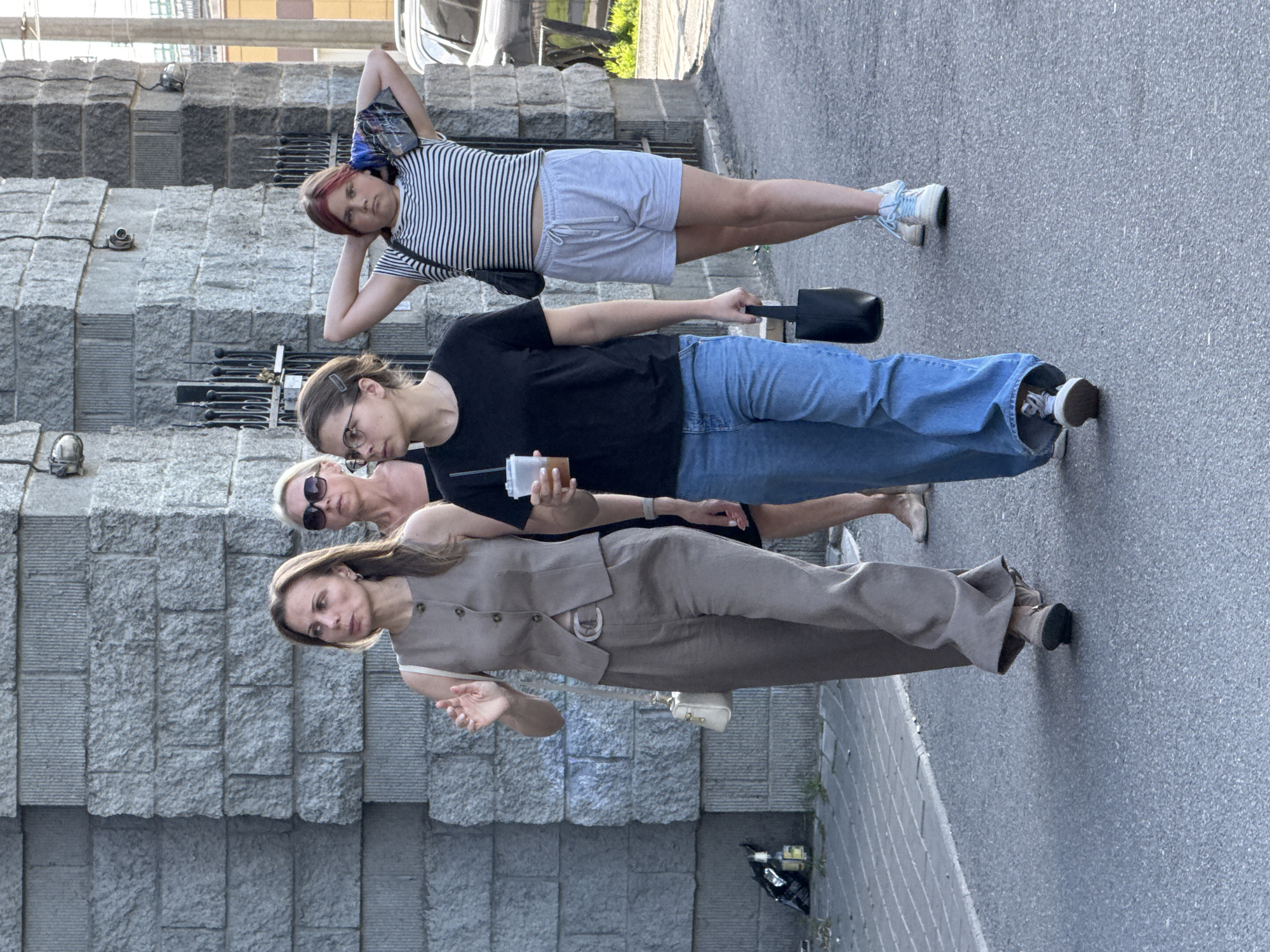 Four individuals, three women and one young person, are walking on a paved street in front of a building constructed of large, rough-hewn grey stone blocks. The woman in the foreground on the left is wearing a light brown sleeveless vest, a white top, and matching light brown pants with a wide belt. She is looking to her left and has her right hand raised slightly, as if waving or gesturing. She is carrying a small, cream-colored handbag on her left shoulder. Next to her, slightly behind, is a woman wearing sunglasses and a dark top, partially obscured. To the right of the first woman is a young person wearing glasses, a black t-shirt, and blue jeans. They are holding a clear plastic cup with a straw in their right hand and carrying a small black handbag in their left hand. The youngest person, on the far right, is wearing a black and white striped t-shirt and light grey shorts. They have their hands behind their head, holding a folded dark fabric item. This individual has reddish-pink colored hair. The building behind them appears to be a modern structure with visible security cameras and metal bars on windows. The ground in front of the building is a grey paved surface with a contrasting lighter grey striped section to the left. The overall scene suggests a daytime urban environment. This location is believed to be in Saint Petersburg, Russia, given the architectural style and typical urban setting.