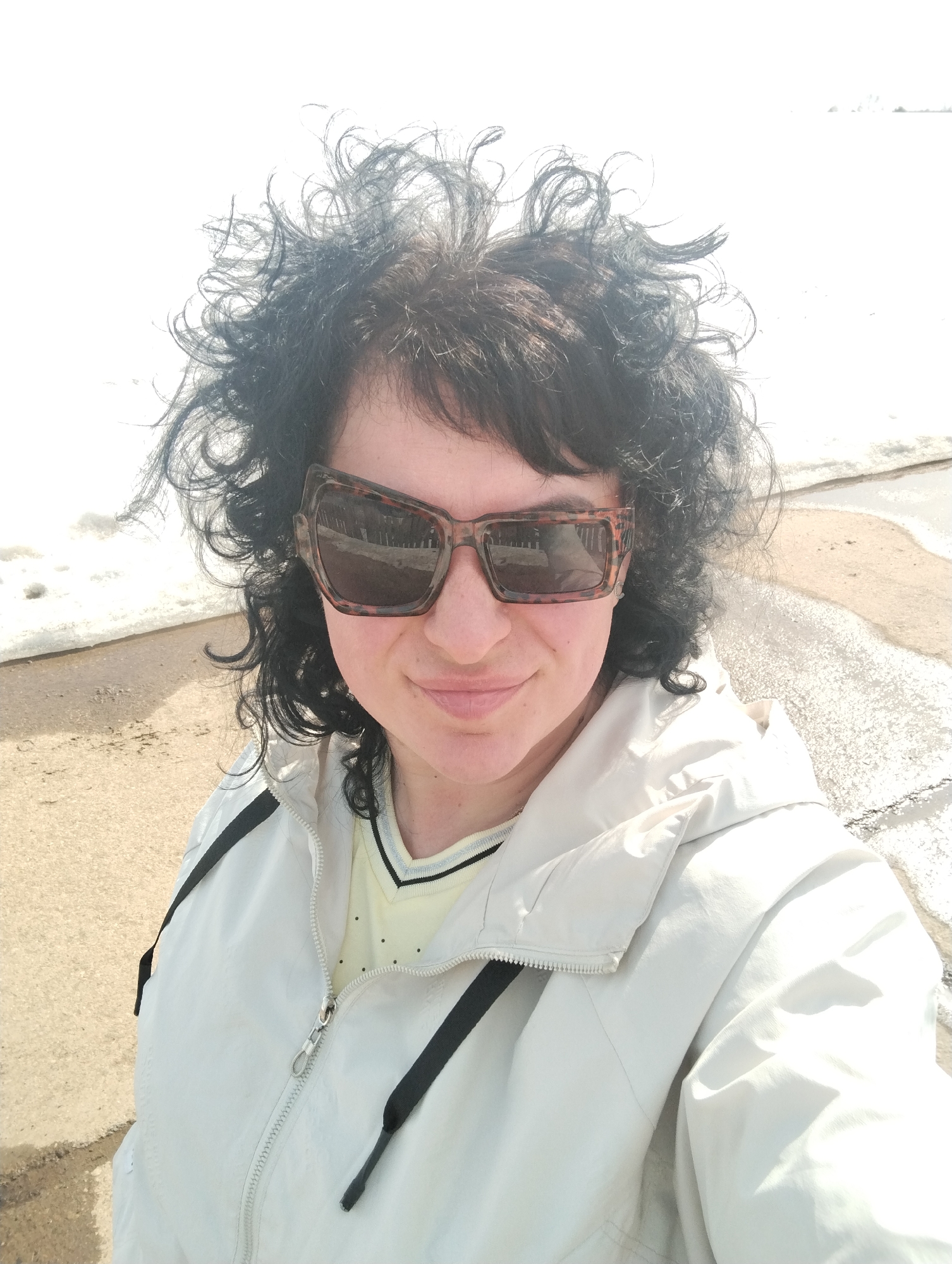 A woman with dark, curly hair and sunglasses takes a selfie. She is wearing a light-colored jacket with black drawstrings. The background shows a partially snow-covered area with some bare ground, possibly a beach or shoreline. The location is identified as Grishanki, Russia.