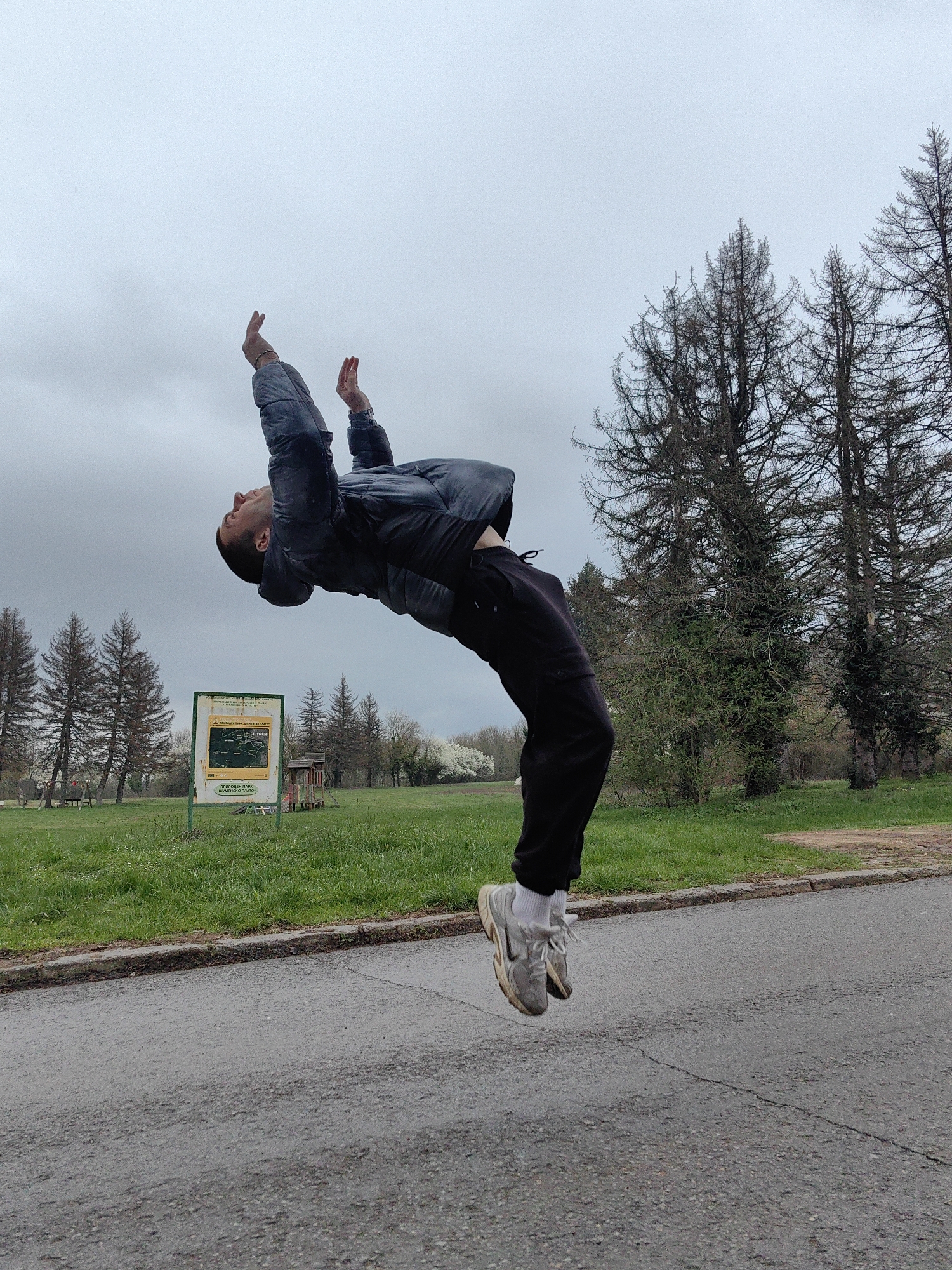 A man is performing a backflip on an asphalt road in a park. He is wearing a dark jacket, black pants, white socks, and gray athletic shoes. His hands are raised above his head. In the background is green grass, bare trees, and a sign in Cyrillic. The scene appears to be outdoors on an overcast day. The location is the Park Priroda "Shumensko Plato" in Shumen, Bulgaria, as indicated by the sign in Cyrillic.