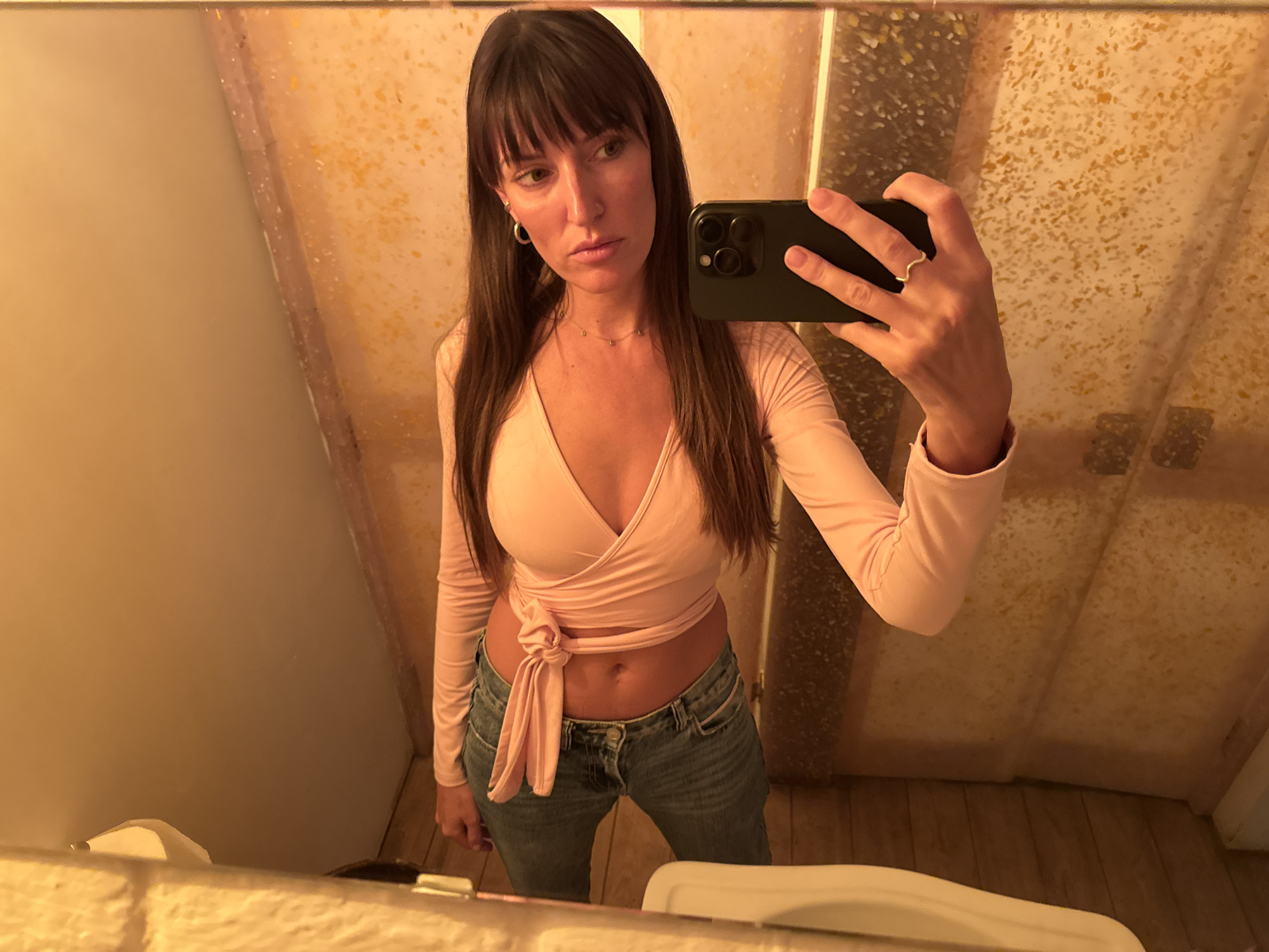 A woman is taking a selfie in a bathroom. She is wearing a light pink wrap top and blue jeans. She has long brown hair and is wearing hoop earrings and a thin necklace. She is also wearing a ring on her right hand. The bathroom has tiled walls and a wooden floor. The lighting is warm and yellowish. The image does not provide any information that would allow for its location to be determined in Moscow, Russia, or any other city.