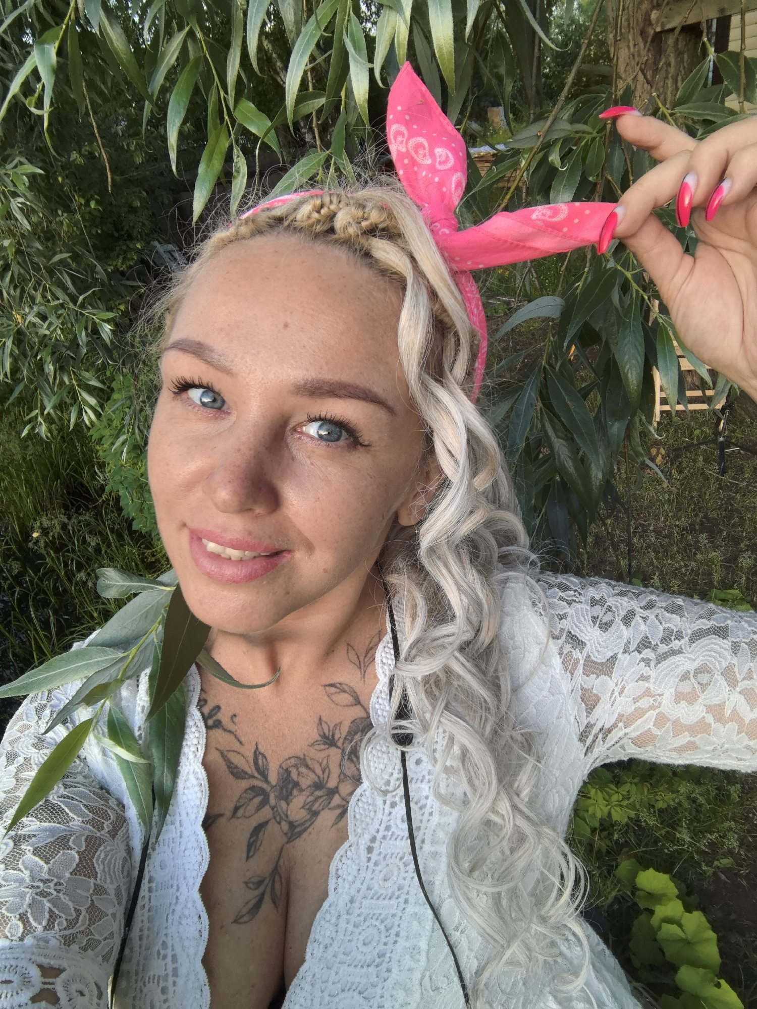 A fair-skinned Caucasian woman with blonde, curly hair and a pink bandana is shown in a close-up outdoor portrait. She is wearing a white lace top and has a tattoo on her chest. Her eyes are blue and she is smiling. Her right hand is raised, with long, bright pink acrylic nails, and she is touching her hair. The background is filled with green foliage, suggesting a natural, possibly park or garden setting. The lighting suggests it is daytime. The image does not provide any specific details to identify the location as Noviy, Russia.
