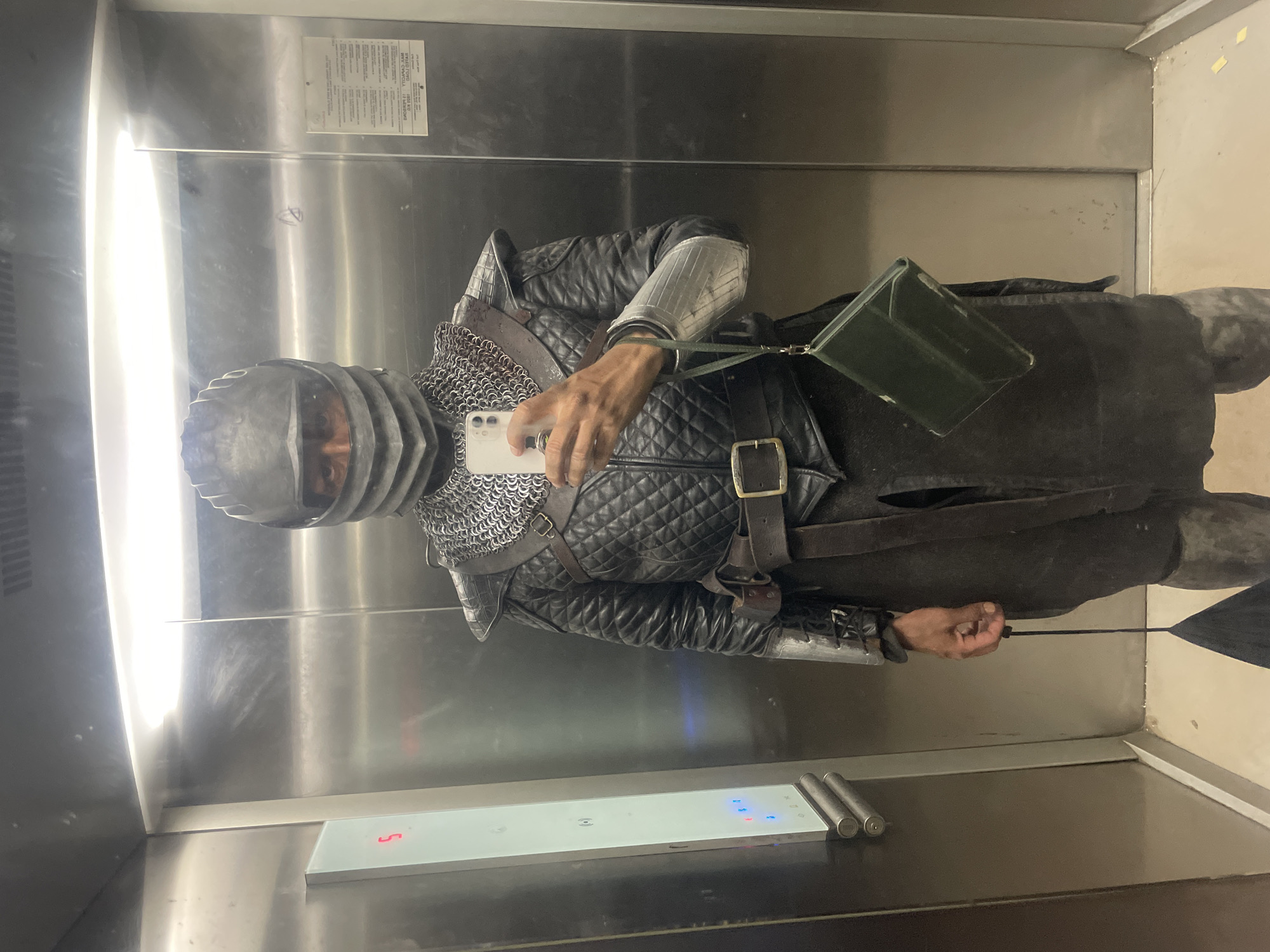 A person wearing a full suit of medieval armor is taking a selfie in an elevator. The armor is primarily black leather with chainmail and a helmet. The person is holding a smartphone in their right hand. There is a green shoulder bag slung across their body. The elevator has stainless steel walls and a control panel with buttons. There is an information sticker on the wall. The location is Unknown, Unknown.