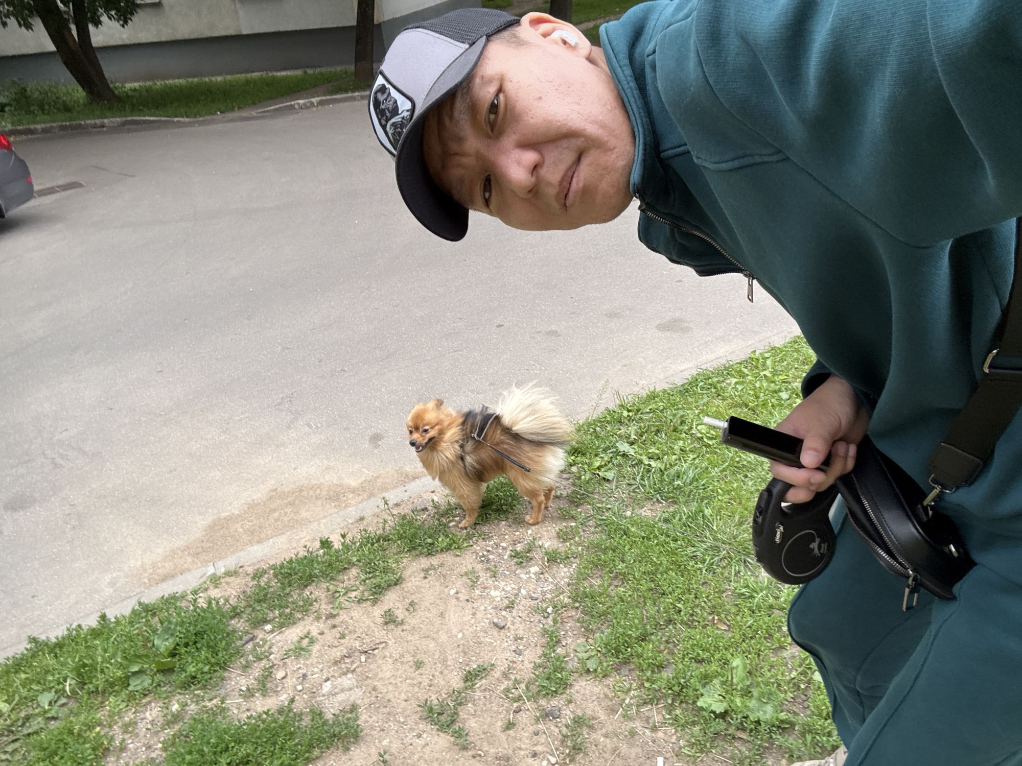 A person wearing a teal tracksuit and a grey and black baseball cap is taking a selfie outdoors. The person is holding a black object, possibly a vape pen, in their right hand, and a black retractable dog leash in their left hand. A small, fluffy, tan Pomeranian dog is standing on a grassy area to the left of the person. The background consists of a paved road, a sidewalk with grass and dirt, and a building with grey siding. The overall scene appears to be a residential street or park area in Moscow, Russia.