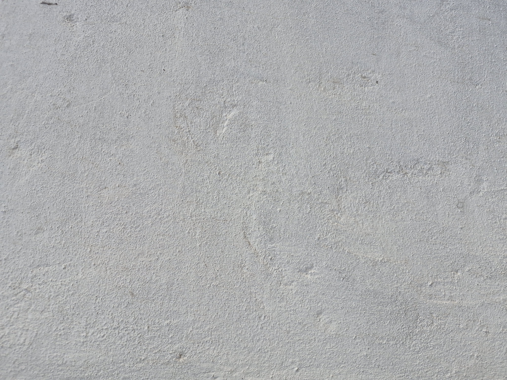This is a close-up, overhead shot of a lightly textured, pale gray surface. The texture appears granular, suggesting it could be concrete, plaster, or a similar material. There are subtle variations in shade and some faint, irregular marks or smudges across the surface. In the top left corner, a small, dark, elongated object or shadow is visible. The lighting suggests it is daytime, and the overall impression is of a mundane, outdoor surface, possibly a wall or pavement. No people, objects, or text are clearly discernible. The context provided indicates the location is in Қанабек, Kazakhstan.