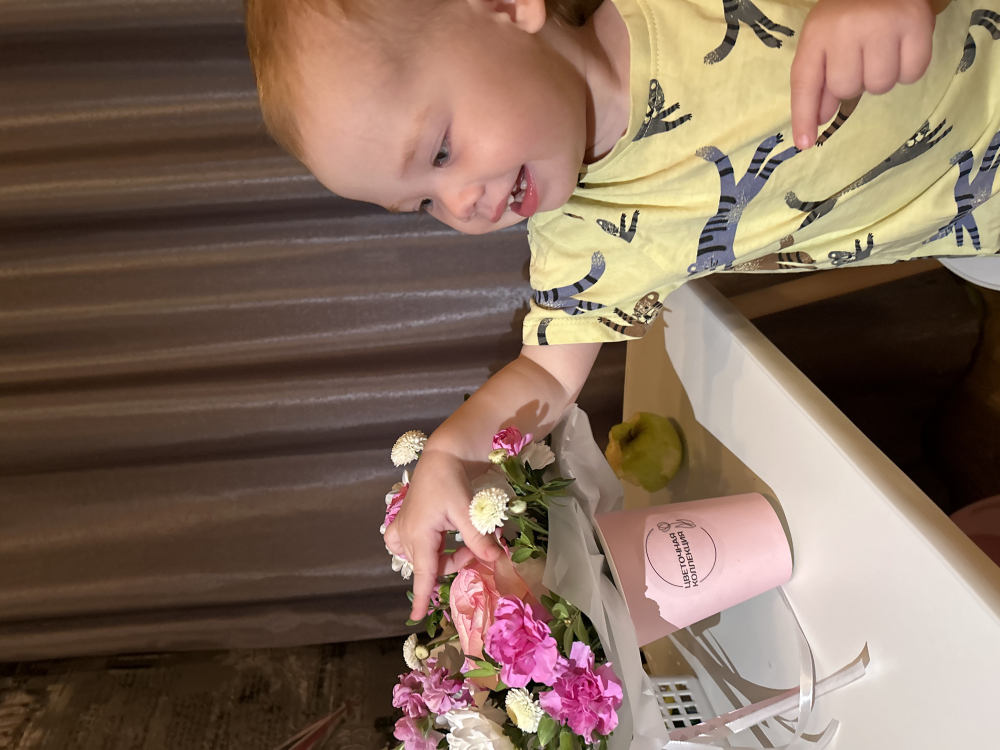 A child in a yellow t-shirt with cat designs is reaching for a bouquet of flowers in a pink container. A green apple sits nearby on a white surface. The image was taken in Chaikovsky, Russia.