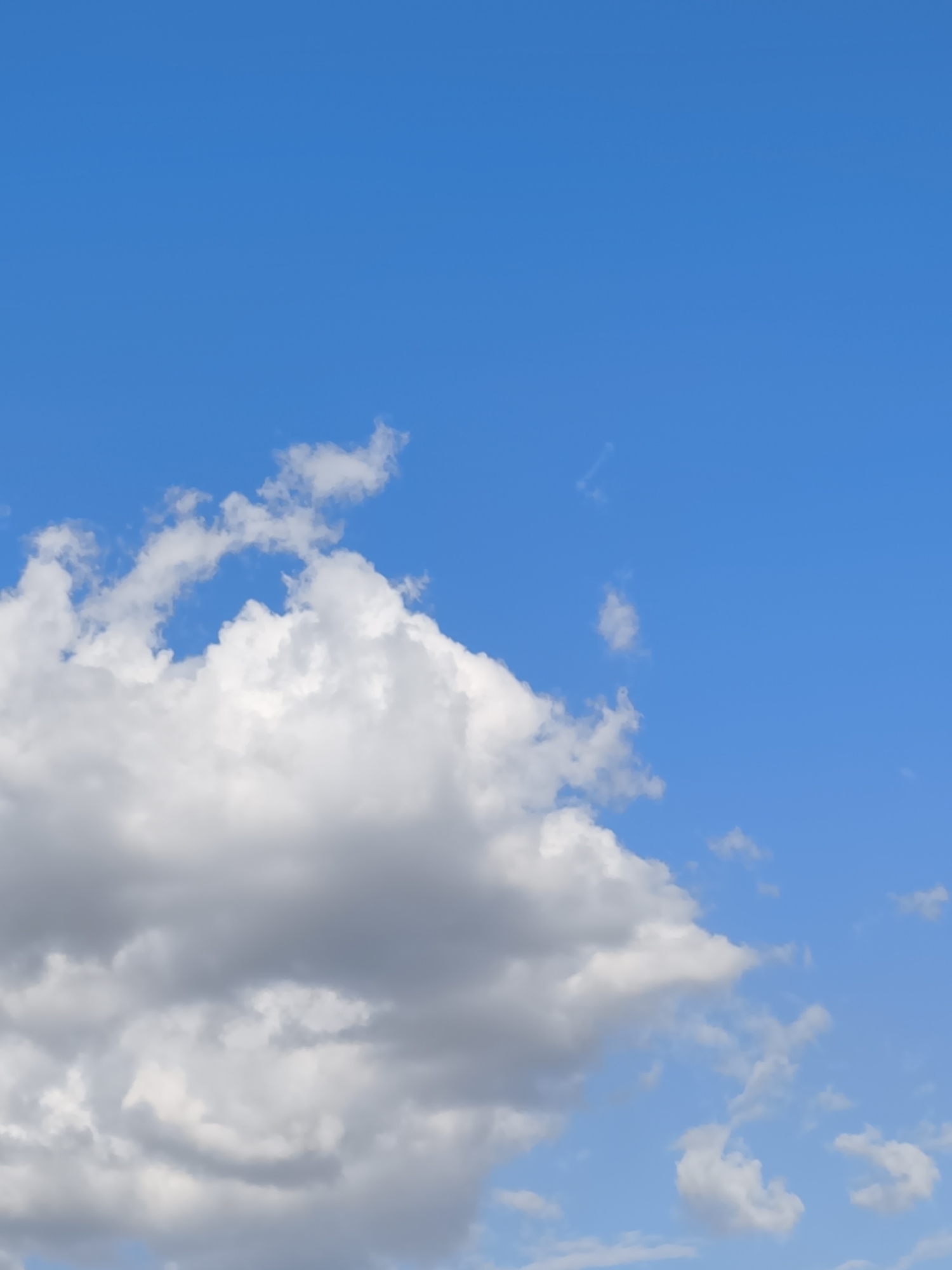 The image is a photograph of a bright blue sky with scattered white and grey clouds. The clouds are primarily located in the lower half of the image, with one large, fluffy cloud dominating the left side. The sky is clear and appears to be taken on a sunny day. There are no people, buildings, or other identifiable objects in the image. The photograph does not provide any specific information or visual cues that would indicate its location within Chișinău, Moldova.