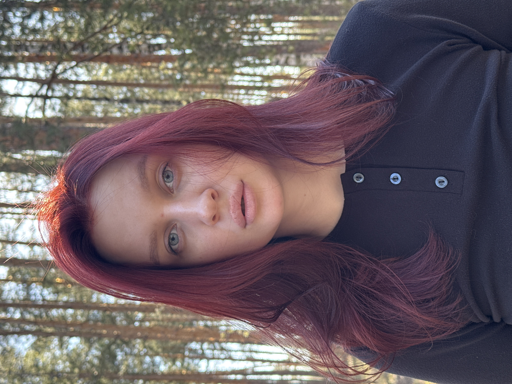 A young woman with dyed red hair and light eyes is centered in the image. She wears a black long-sleeved shirt with small buttons. The background is blurred but appears to be a forest. The image was taken in an outdoor setting with natural lighting. There is no indication the photo was taken in Nizhny Novgorod, Russia.