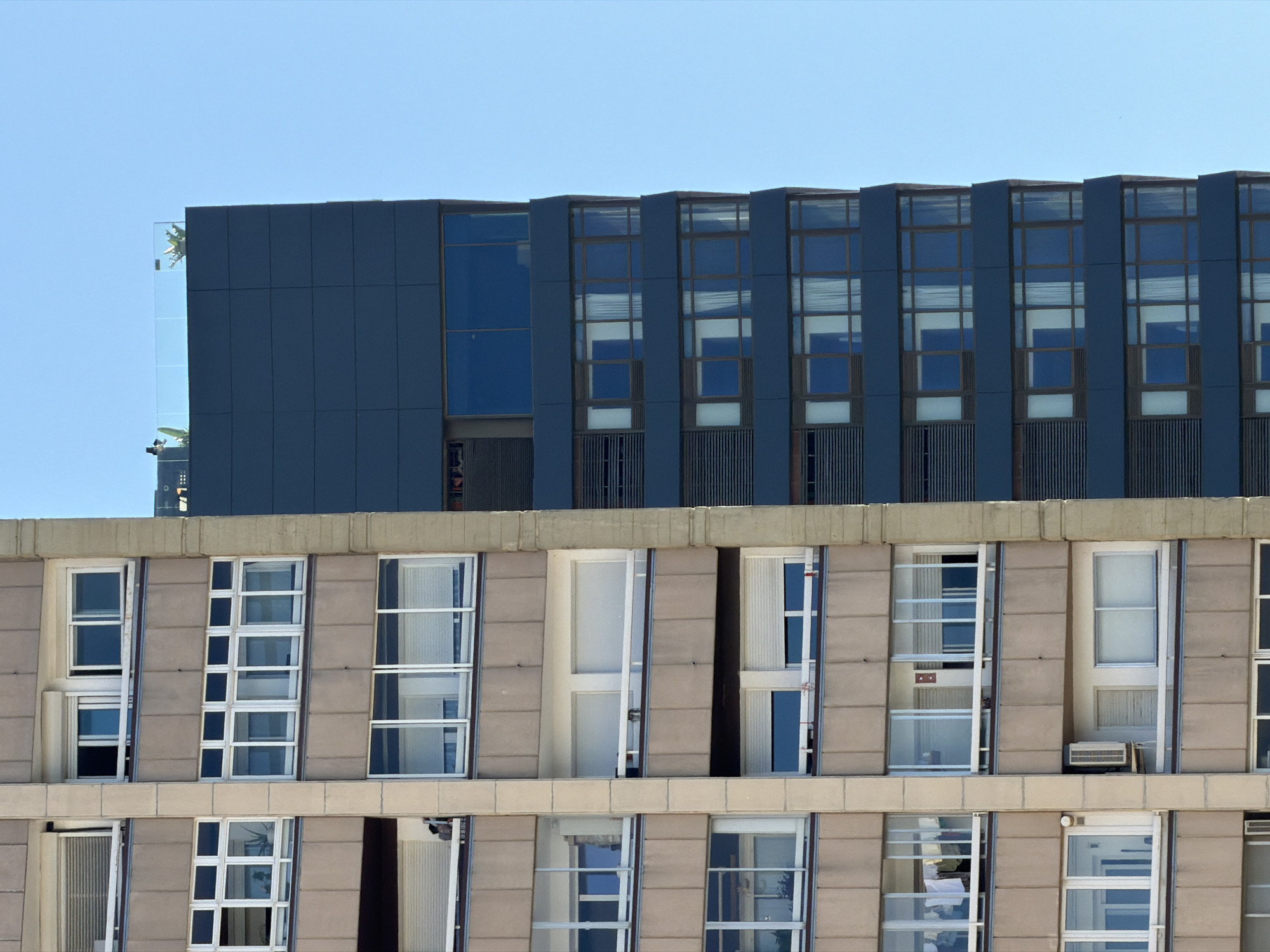 This image shows the facades of two buildings, one behind the other, against a clear blue sky. The building in the foreground has a light beige facade with horizontal paneling and multiple rows of windows. These windows are mostly white with multiple panes and appear to be open in some cases, revealing the interiors of rooms. Some windows have what look like blinds or screens. There are air conditioning units visible in some of the lower windows. The building behind is darker, with a deep blue facade. This building features a repeating pattern of tall, narrow windows with dark frames, divided by vertical blue panels. Some of these windows appear to be filled with darker blue glass, reflecting the sky, while others have lighter sections, possibly indicating internal lighting or different materials. A thin, reflective glass structure is visible at the top left edge of the darker building, with some green foliage or decorative elements visible through it. The overall scene suggests an urban setting, possibly in Barcelona given the architectural style and the clear weather, indicative of a sunny day. No people are visible in the image.
