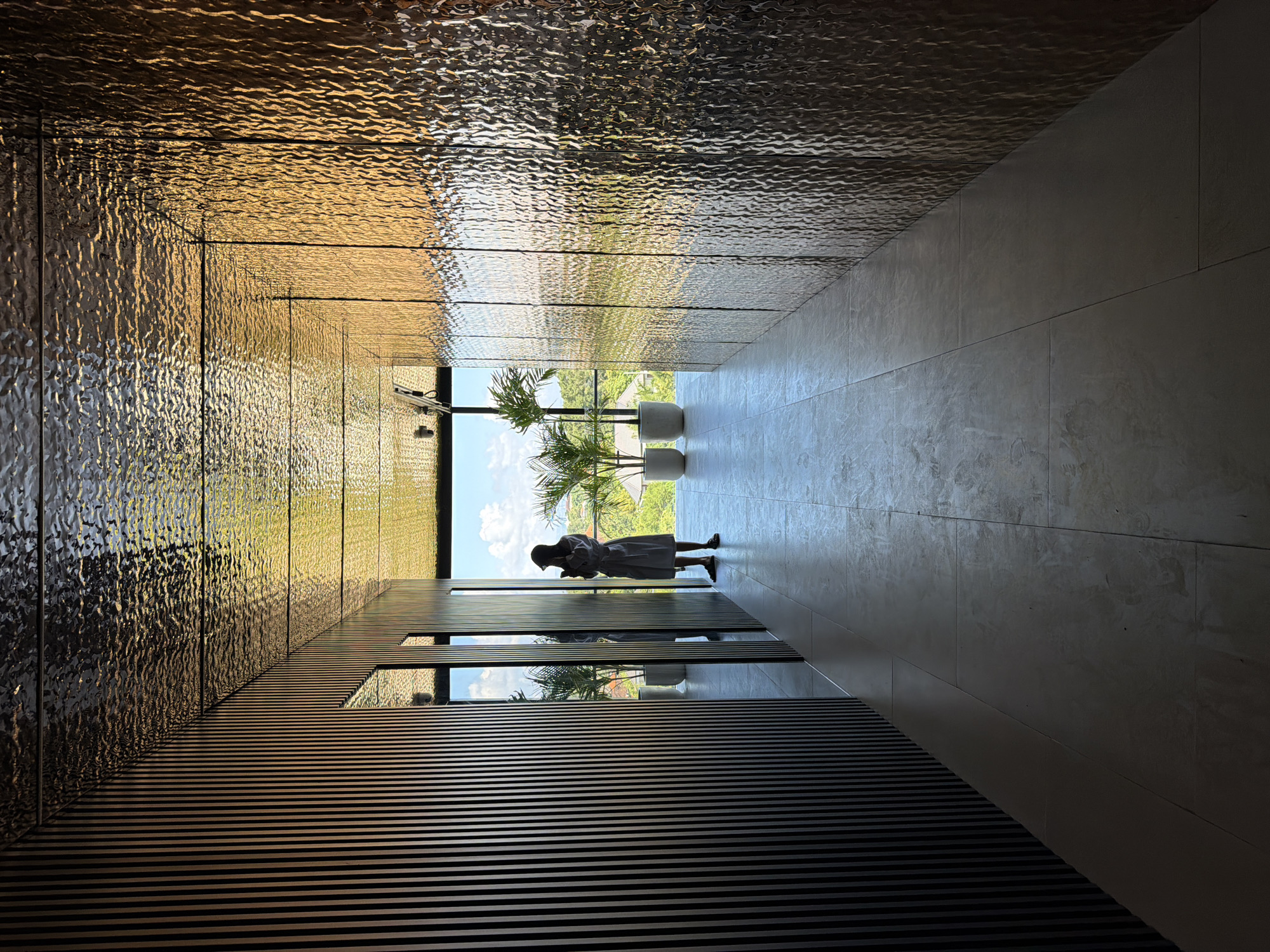 The image captures a modern architectural hallway or corridor in North Kuta, Indonesia, looking towards a bright outdoor scene. The composition is striking, featuring strong contrasts in texture, light, and shadow.
The left side of the hallway, functioning as a wall or ceiling, is covered in highly textured, reflective panels that shimmer with golden and bronze light, reflecting distorted views of the surroundings. This creates an effect reminiscent of rippling water. In contrast, the right side, forming the floor and an opposing wall, consists of smooth, light-toned panels, giving a clean and minimalist aesthetic. Along the lower left side, a dark, horizontal slatted structure runs parallel to the floor, adding a repeating linear pattern.
At the end of this perspective-driven corridor stands a person, silhouetted against the brilliant outdoor light. The individual, appearing to be a woman wearing a hat and a long dress or robe, is seen from behind, looking downwards, possibly at a device or absorbed in thought. Flanking her are two potted palm plants, positioned at the threshold of the expansive opening that leads to the exterior.
Beyond the hallway, a vibrant outdoor landscape is visible, characterized by lush green foliage under a clear blue sky, indicating a sunny daytime setting. The strong backlighting emphasizes the silhouette of the person and the reflective qualities of the architectural materials. The scene evokes a sense of calm and contemplation. There is no visible text in the image.