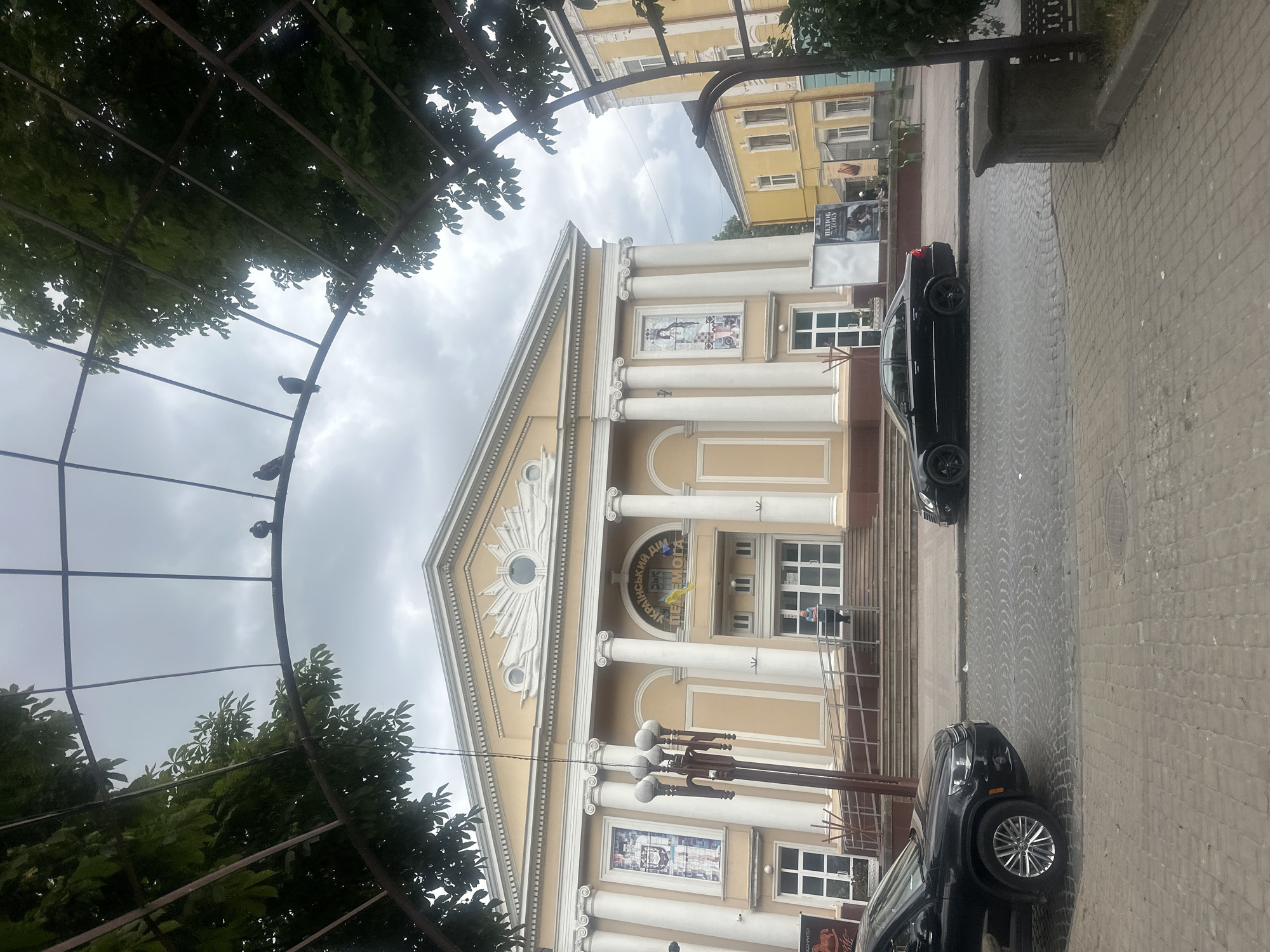 In Ternopil, Ukraine, a black SUV and a black sedan are parked on a cobblestone street in front of a large, cream-colored building with neoclassical architecture. The building has a grand entrance with white columns and ornate detailing. Above the entrance, a sign reads "Український дім" (Ukrainian House) and "ПЕРЕМОГА" (Victory) with a Ukrainian flag. There are posters displayed on the building's facade. In the foreground, a metal archway covered with green leaves frames the scene. Several pigeons are perched on the archway. The sky is overcast.