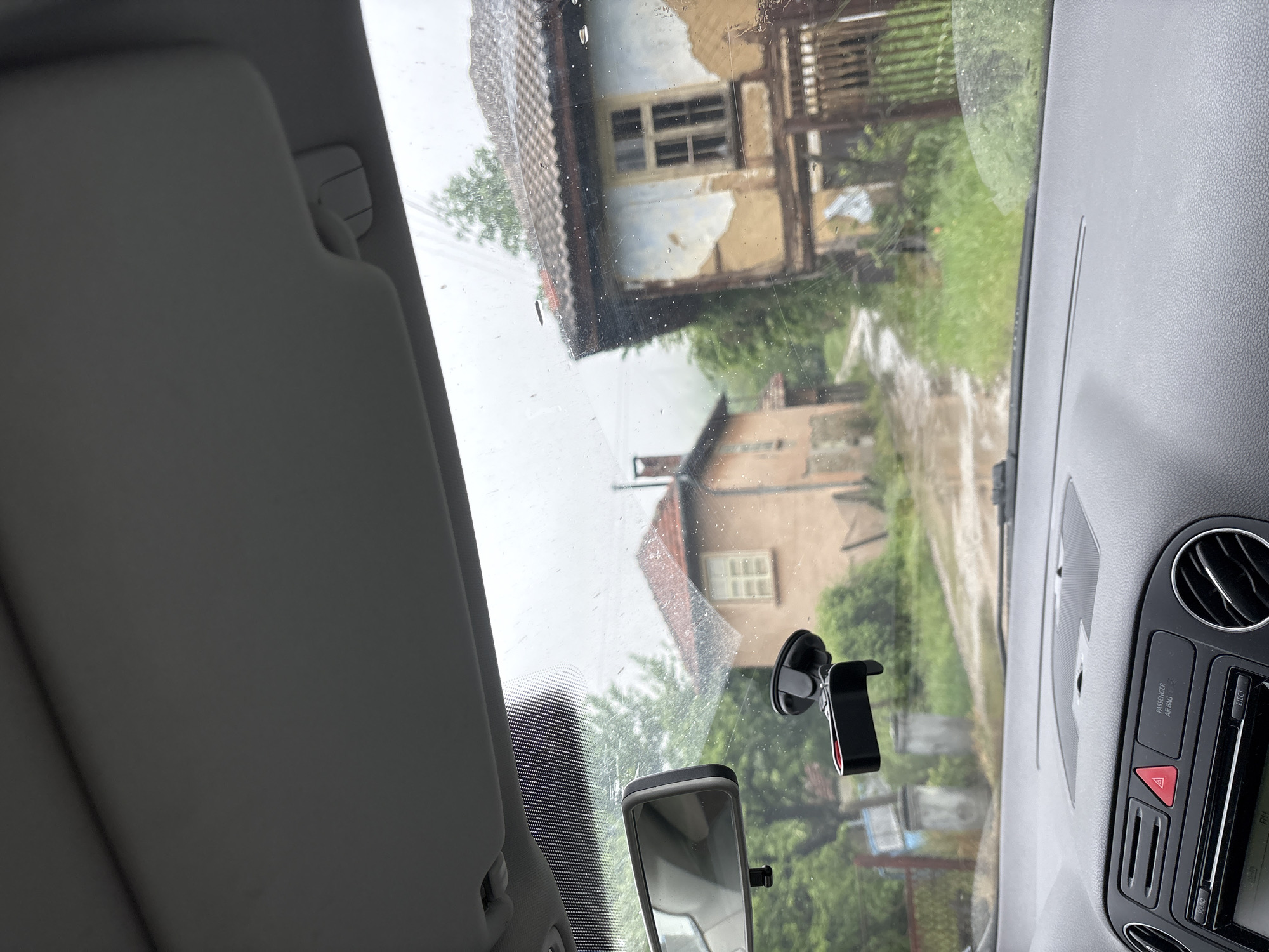 The image shows a view from inside a car, looking through the windshield on a rainy day. Two buildings are visible, suggesting a residential area. The location is Plachkovtsi, Bulgaria, based on the prompt. The building directly visible through the windshield appears to be older, perhaps partially dilapidated, with visible windows and a partial view of its roof. A second building is visible farther back with a red roof. In the foreground of the interior, a car's dashboard is partially visible along with an interior mirror and a cell phone mount on the windshield. The sun visor is partially down on the left side of the frame.