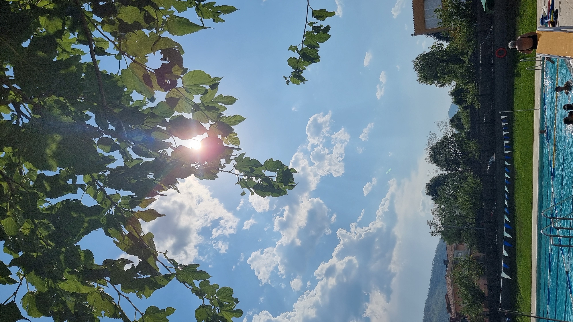 The image depicts an outdoor swimming pool area on a sunny day, with lush green trees and a bright blue sky filled with white, fluffy clouds dominating the upper portion. The sun is visible through the leaves of a tree in the foreground, casting lens flare. In the distance, a town or village with terracotta-roofed buildings and rolling hills can be seen. The swimming pool is clean and blue, with lane markers visible. Several people are in or around the pool, including a woman in a black swimsuit sitting on the edge of a yellow slide, and others swimming. The scene suggests a recreational or public space, likely a municipal pool in Sant Joan Les Fonts, Spain, given the architectural style and the surrounding landscape which is characteristic of the region.