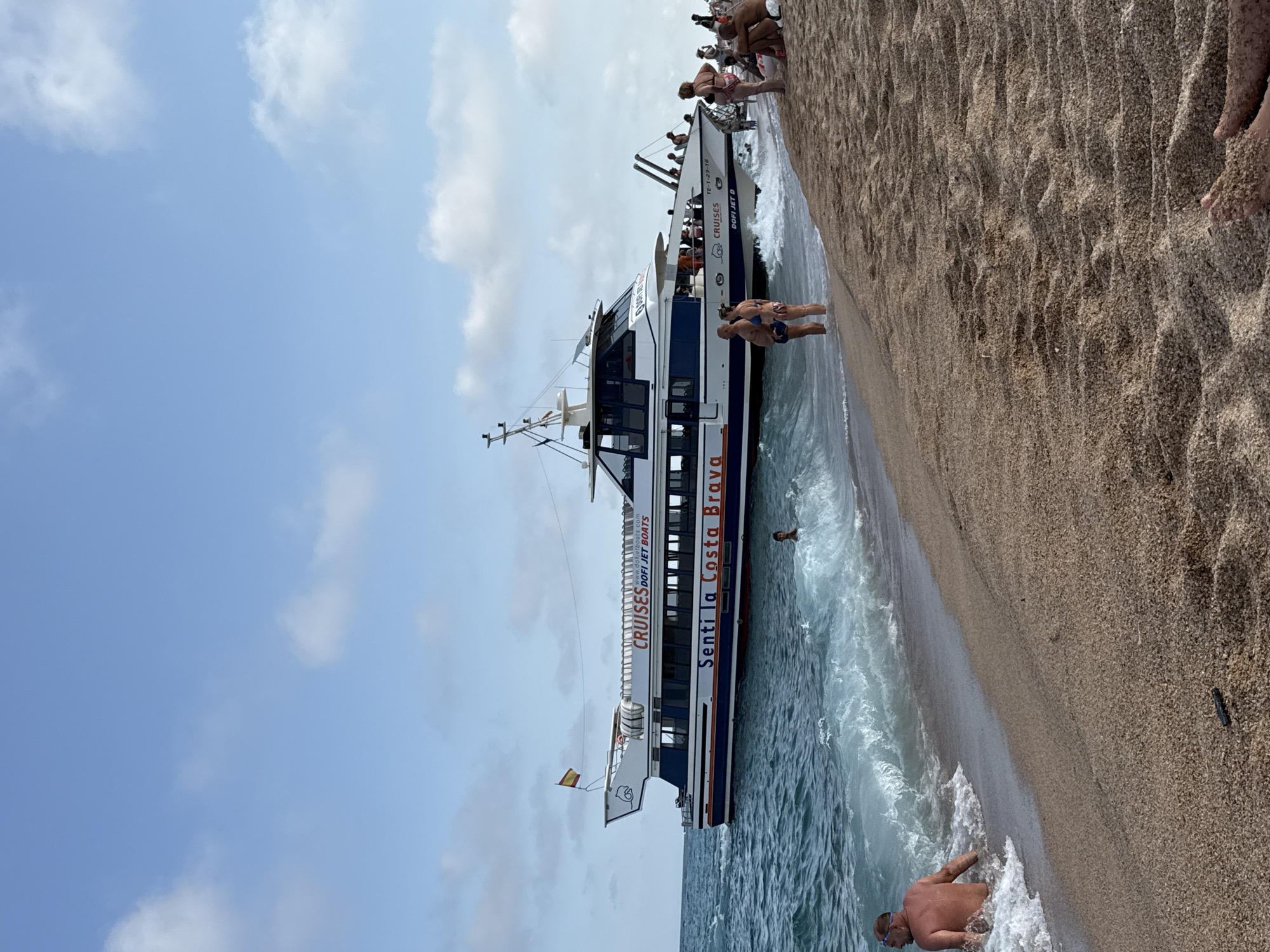 The image depicts a sunny day at a beach in Blanes, Spain. A large passenger boat, "CRUISES Senti la Costa Brava," is anchored near the shore. The boat is white with blue and orange trim, and it appears to be carrying passengers. A Spanish flag is visible on the boat's mast. Several people are on the beach, some wading in the water, while others are sitting or standing on the sand. The sand is light brown and appears to be fine-grained. The water is a clear blue-green, with gentle waves breaking on the shore. The sky is a light blue with scattered white clouds. In the foreground, a man is partially submerged in the water, wearing swimming goggles. Further down the beach, a group of people are gathered. The overall scene suggests a leisurely day at the beach.