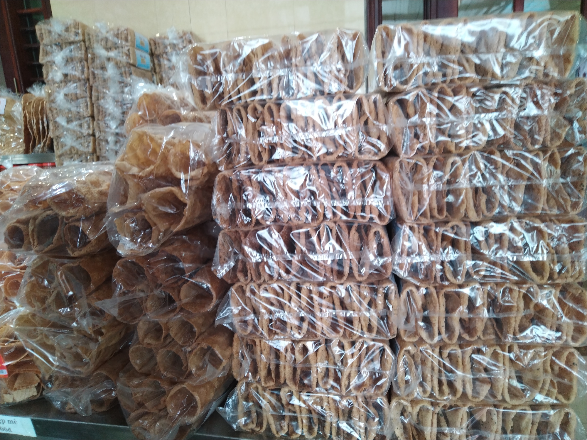 The image shows stacks of packaged snacks. On the left, there are rolls of what appear to be wafers or crepes. To the right, there are neat stacks of rectangular packages containing rows of a tubular, lattice-like snack. All items are individually wrapped in clear plastic. There are more stacks of packaged snacks visible in the background. The location is Chu Văn An Ward, Vietnam.