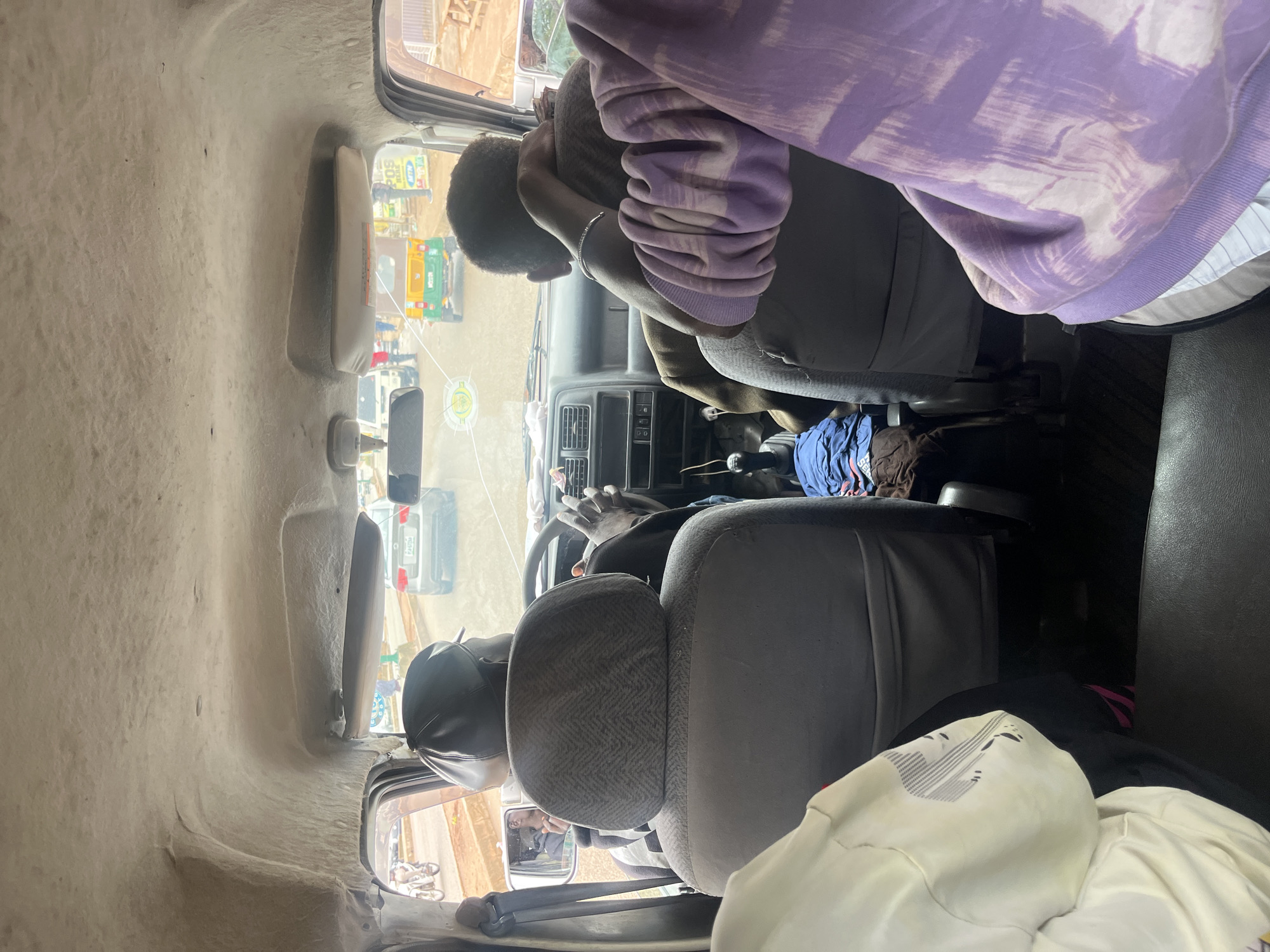 The image captures the interior of a moving vehicle, likely a van or a bus, on a street in Bukuru, Nigeria. The perspective is from the back seat, looking forward. A man in a black cap is driving, his hands on the steering wheel. Next to him, another person is seated, resting their head on their arm. In the foreground, a person wearing a white garment with a patterned design is visible on the left, and another person in a purple tie-dye hoodie is on the right, partially obscuring the view of the driver and the front passenger. The interior of the vehicle appears worn, with textured fabric on the ceiling and upholstered seats. Outside the vehicle, a grey sedan and a green and yellow auto-rickshaw are visible on the road, along with other indistinct figures and vehicles, suggesting a busy urban environment. The lighting suggests it is daytime.