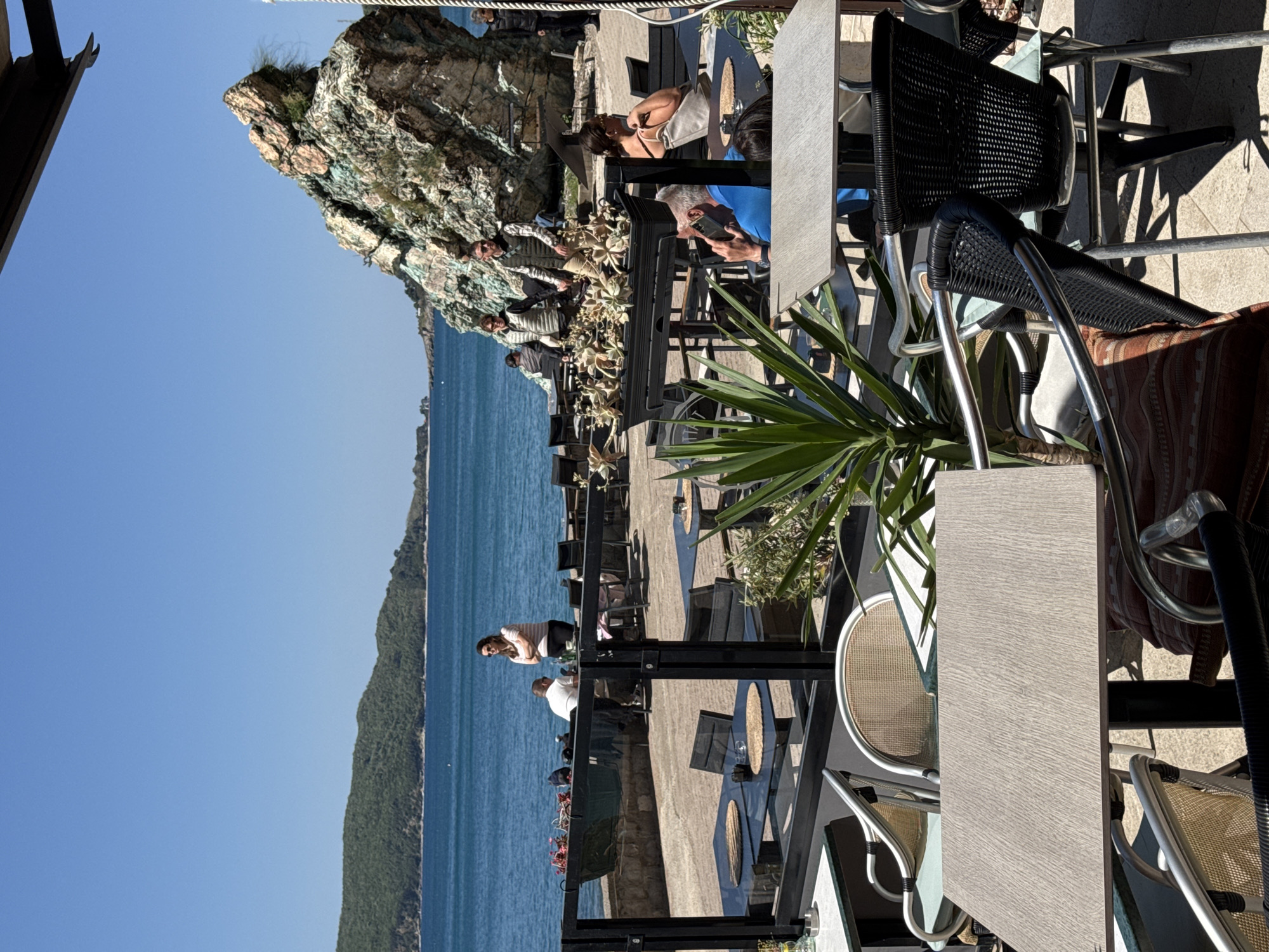 This image captures an outdoor cafe or restaurant overlooking the sea in Rafailovići, Montenegro, under a bright, clear sky. The scene is depicted as if viewed from an elevated position, looking down and across the establishment.
In the mid-ground, a prominent, rugged rocky cliff covered with sparse vegetation rises dramatically. Below and to its left, the calm, blue-green expanse of the Adriatic Sea stretches out, meeting a distant, forested coastline under a clear horizon.
The cafe area features numerous outdoor tables and chairs arranged on a paved surface. In the immediate foreground, a light-colored rectangular table and a black mesh chair with a metallic frame are visible, accompanied by a large green plant, possibly a yucca, partially obscuring the view. Further into the cafe, several individuals are seated or standing. One man in a blue shirt is engrossed in his mobile phone, while a woman in a light-colored top sits at an adjacent table. Near the sea-facing edge, a woman in a striped shirt stands, looking out at the water, and a man in a white shirt is seated nearby. In total, approximately eight to ten people are present, appearing relaxed and enjoying the sunny weather and scenic views.
The tables vary in design; some are dark glass with circular patterns, while others are a lighter wood-like material. Chairs are a mix of black woven mesh and lighter-colored frames with woven seats. Black metal railings and planters with small plants segment the seating areas. The shadows cast by the furniture indicate strong overhead sunlight, suggesting a midday or early afternoon setting. No discernible text is visible within the image.