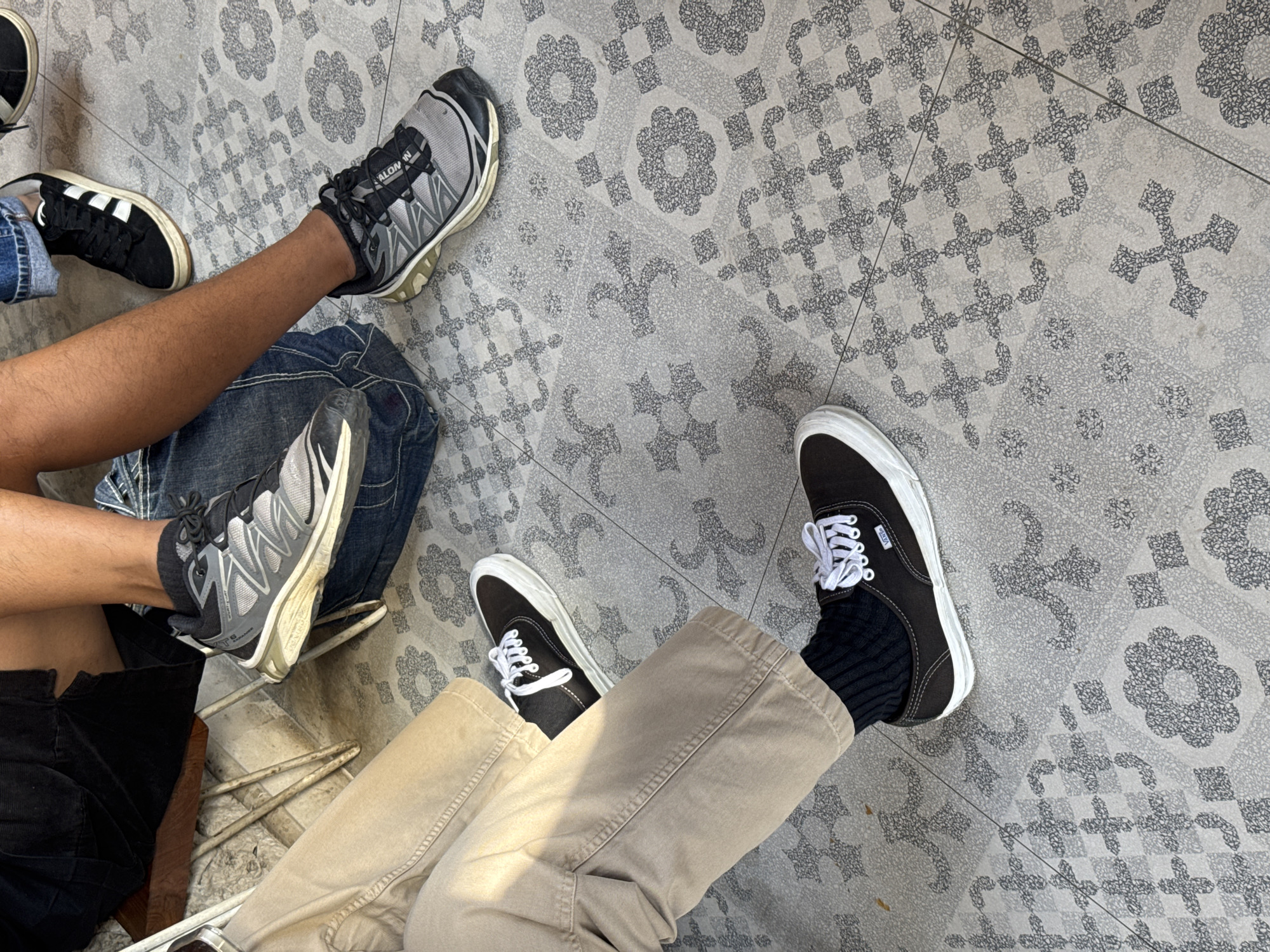 A low-angle, medium shot captures the legs and feet of at least three individuals seated on a patterned tile floor. The tiles are a light grey with a darker grey, ornate floral and cross-like pattern. In the foreground, a person wearing tan pants and black socks is seated with their legs crossed, showing dark brown Vans sneakers with white laces. To the left and slightly behind, another person's legs are visible, wearing tan pants and white sneakers with black laces. Further back and to the right, a person in blue jeans is visible, wearing black sneakers with white accents and laces. One individual's foot is placed on top of a denim-covered surface. The scene appears to be outdoors, possibly in a cafe or public seating area in North Kuta, Indonesia, given the architectural style of the flooring.