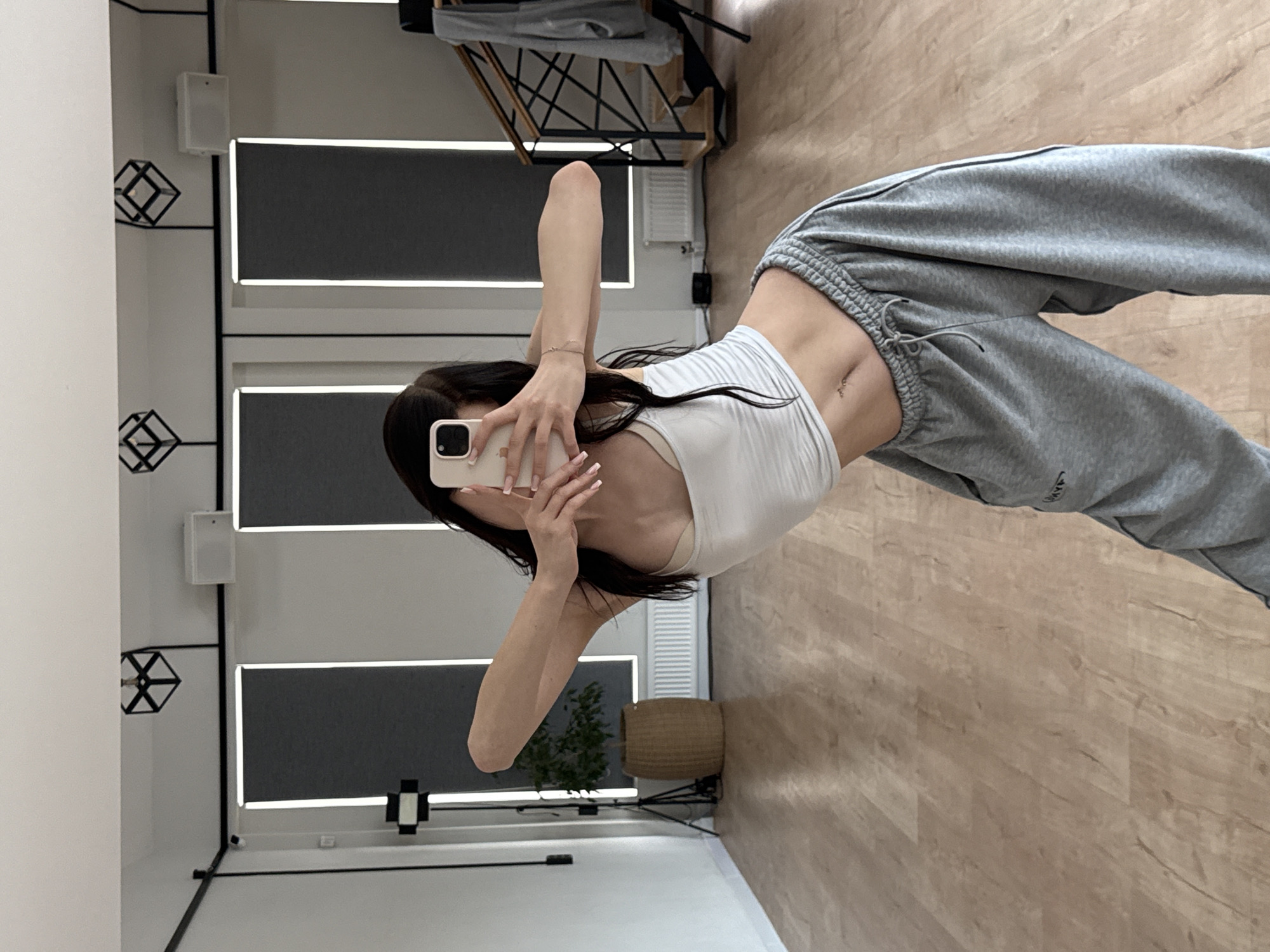 A woman is standing in what appears to be a dance studio with wooden floors and large windows with grey blinds. She is taking a selfie in a mirror, holding a smartphone in her right hand. She is wearing a white crop top and grey sweatpants. Her arms are outstretched, and her body is contorted in a pose. In the background, there is a plant in a woven basket, a tripod with a light, and a staircase with a railing. The lighting in the room is bright. The city of Zakharovo, Russia is not depicted in this image.