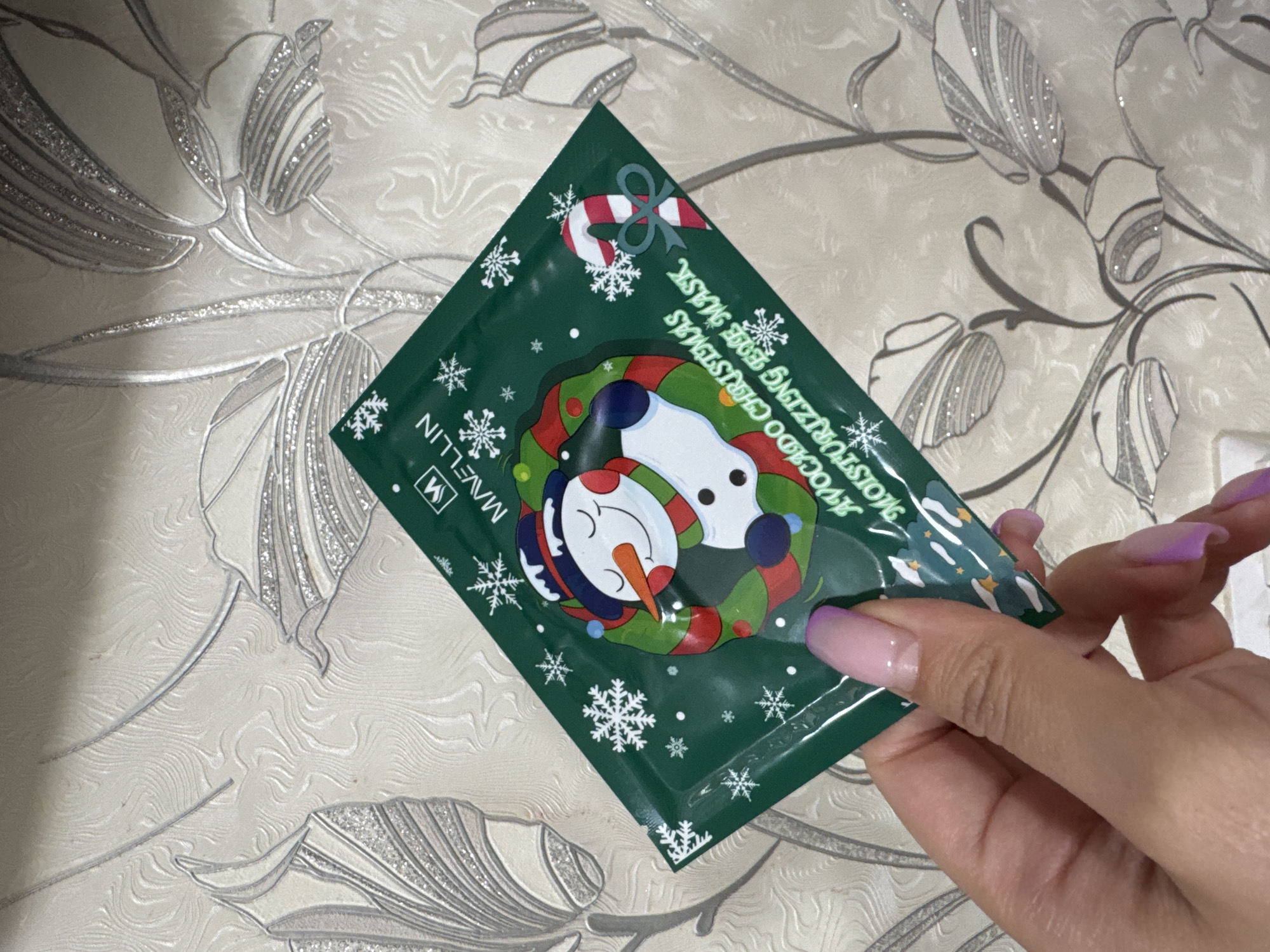 A hand with manicured nails holds a green facial mask packet featuring a snowman and snowflakes design. The packet reads "MAVELLIN AVOCADO CHRISTMAS MOISTURIZING BODY MASK." The background is a beige surface with a subtle floral pattern. The photograph appears to be taken indoors. The location in Nizhnyaya Gar', Russia is not explicitly stated but implied.