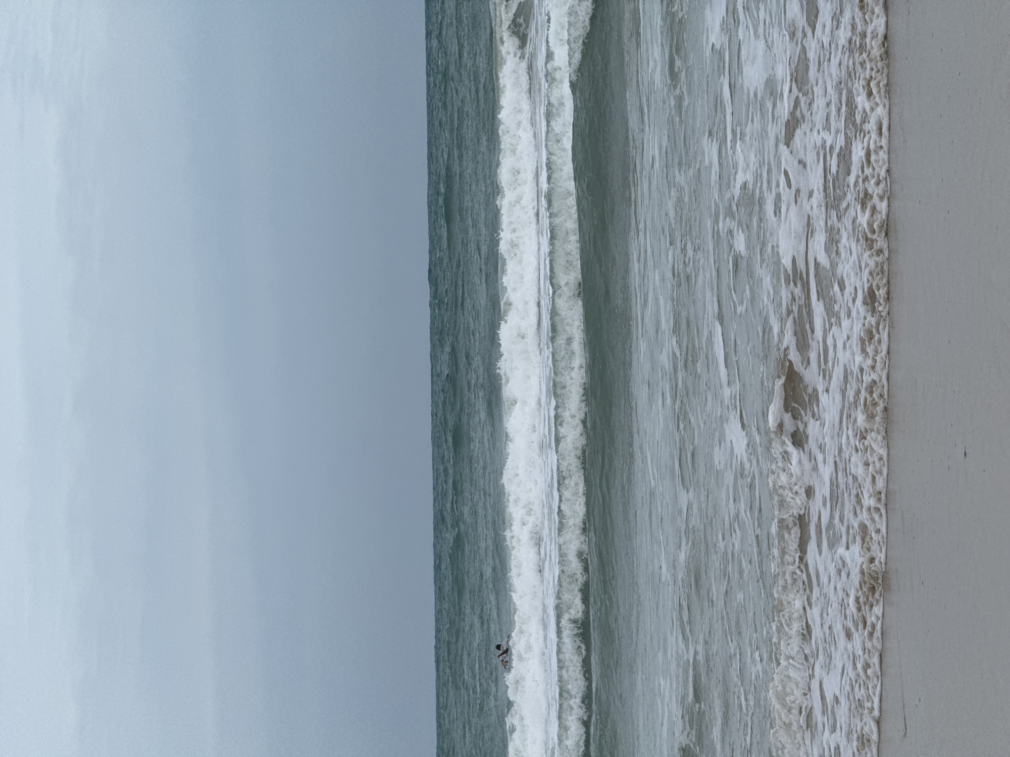 A person is seen swimming in the ocean off the coast of Karon, Thailand. The sky is overcast, and the sea is choppy with moderate waves. The beach in the foreground is sandy and wet from the incoming tide.